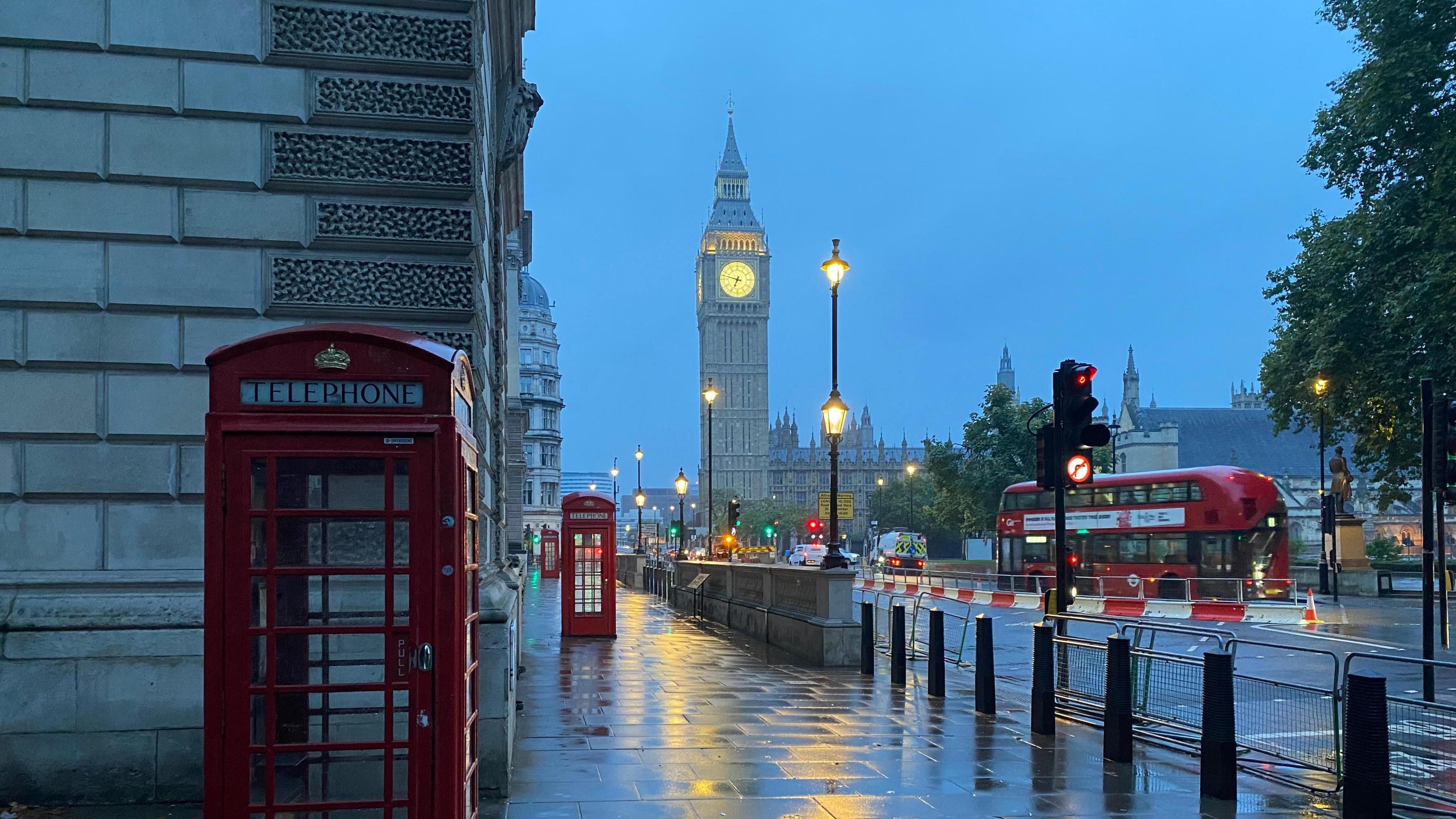 image of a gloomy looking London with overcast skies, some rain on the ground reflecting the light from street lamps.  Red phone boxes and red London bus can been seen