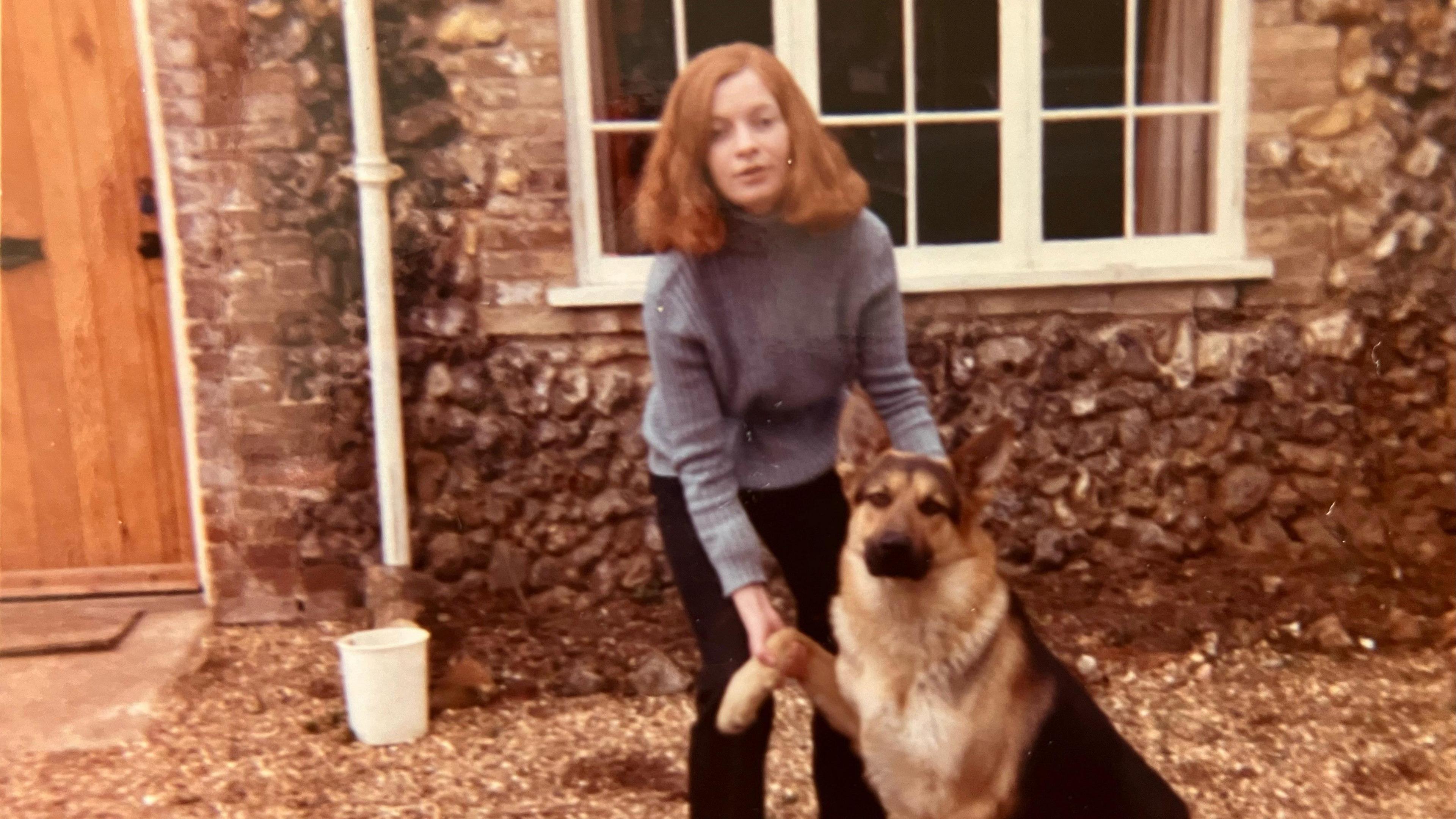 A family photo of Sheenagh, with long, red hair, outside a stone cottage with a German Shepherd dog whose paw she is holding