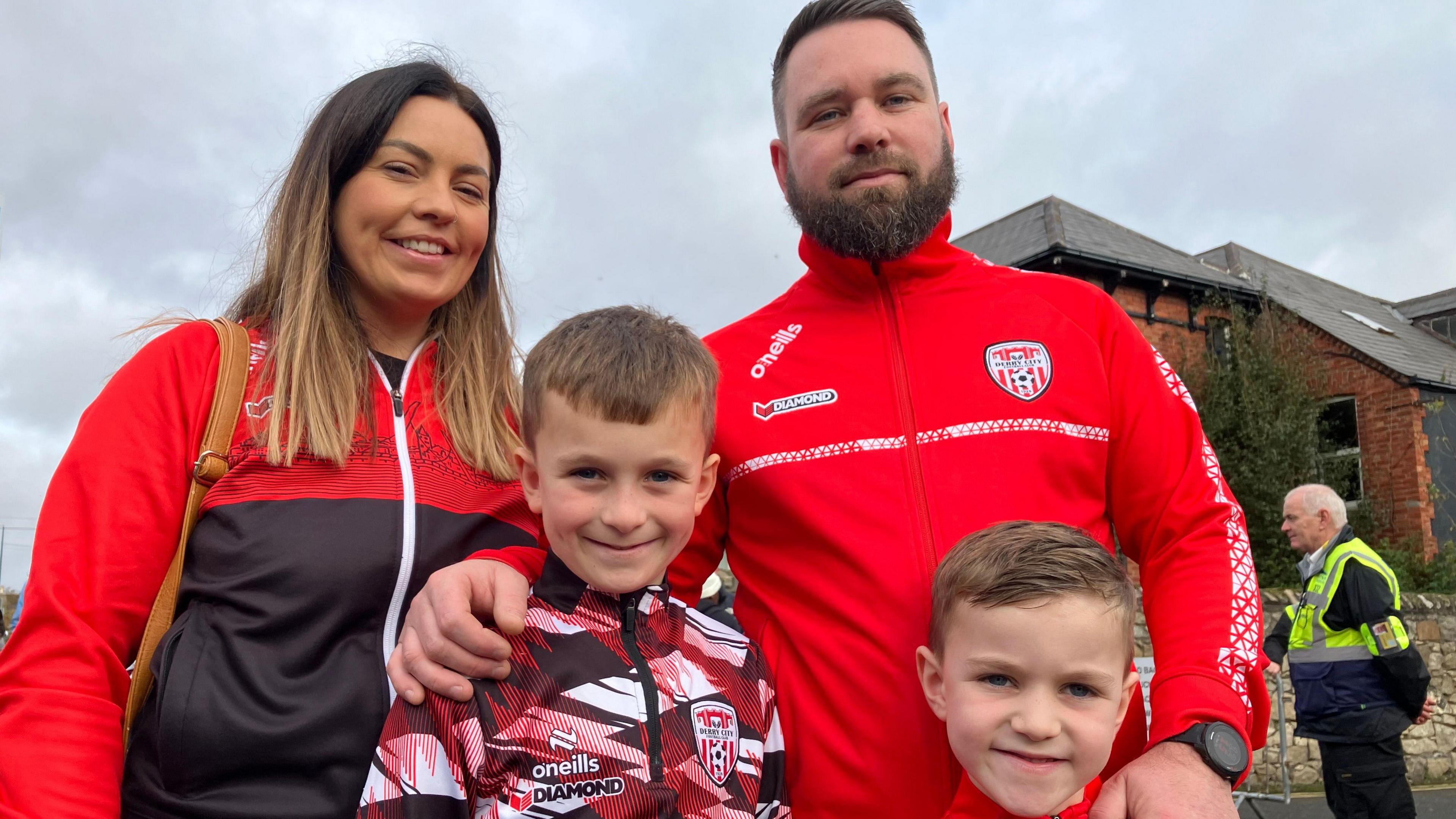 A family of four standing together smiling at the camera. The dad has his arms round his two sons while their mum is standing next to them. They are are wearing the red of Derry City. Behind them a cloudy sky and a house in the background.