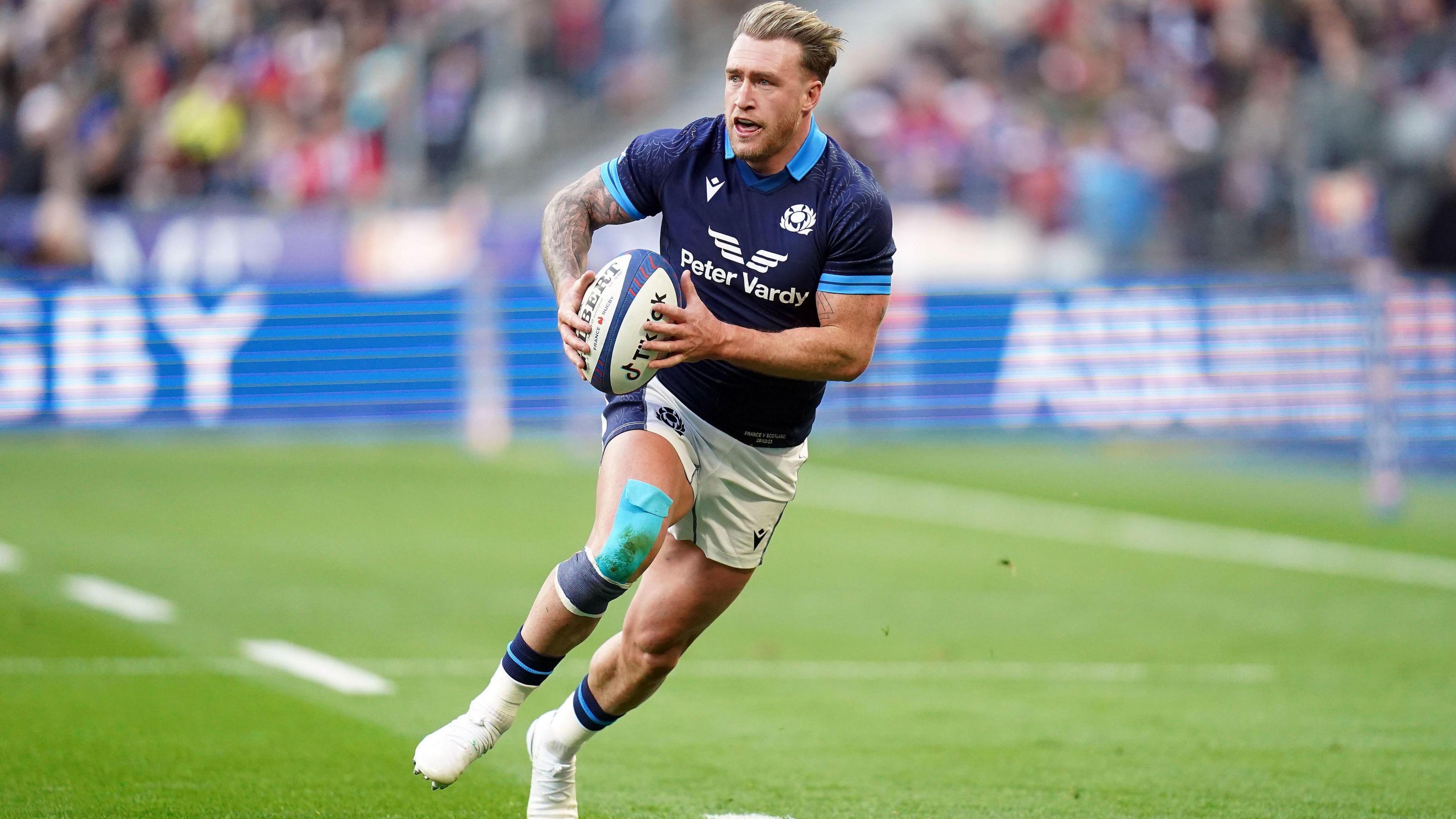 Stuart Hogg in a Scotland rugby top with the ball in hand runs forward with a large bandage on his knee