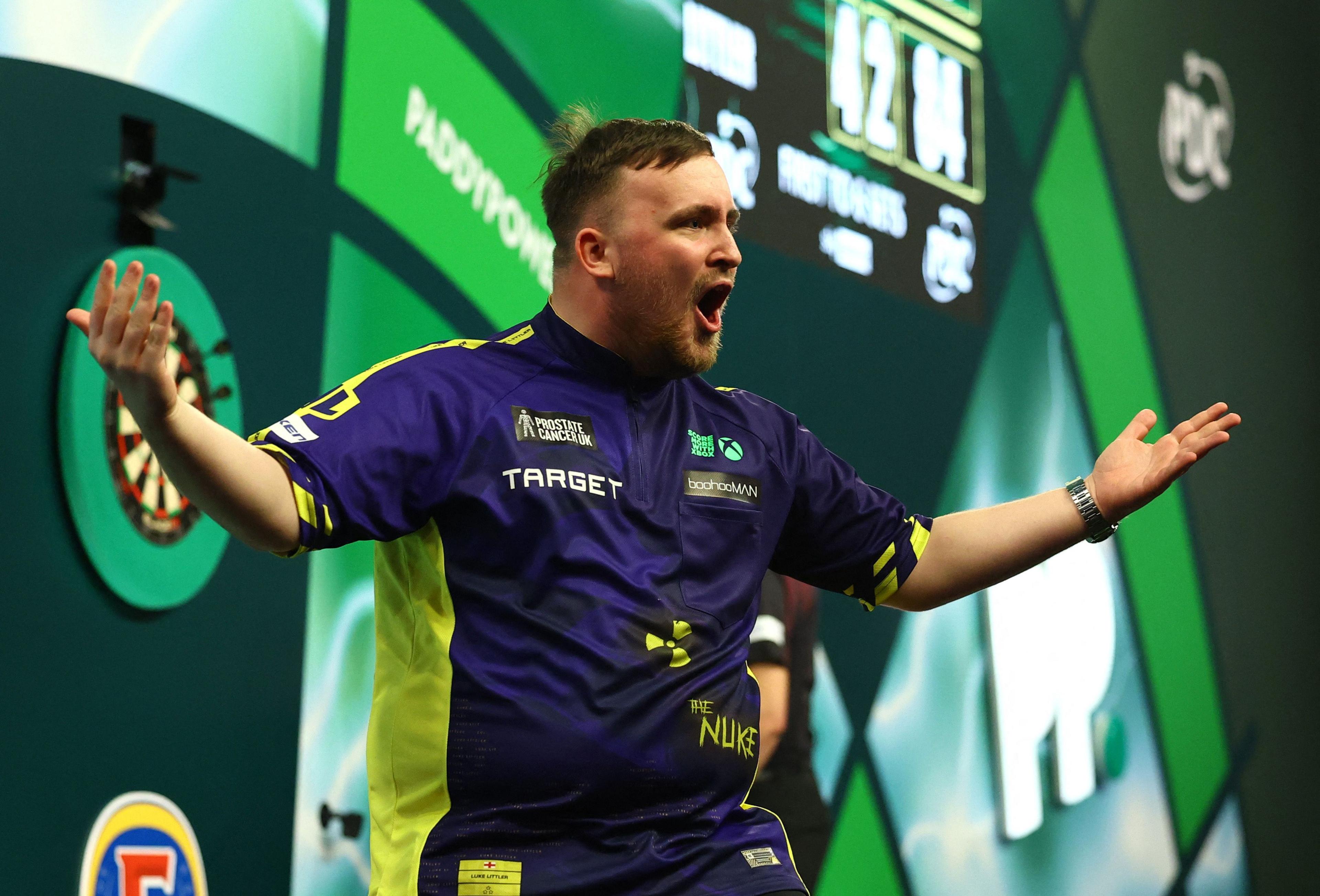 Luke Littler, a young man in a navy and yellow sports top, shouts at the crowd on a stage. There is a darts board behind him