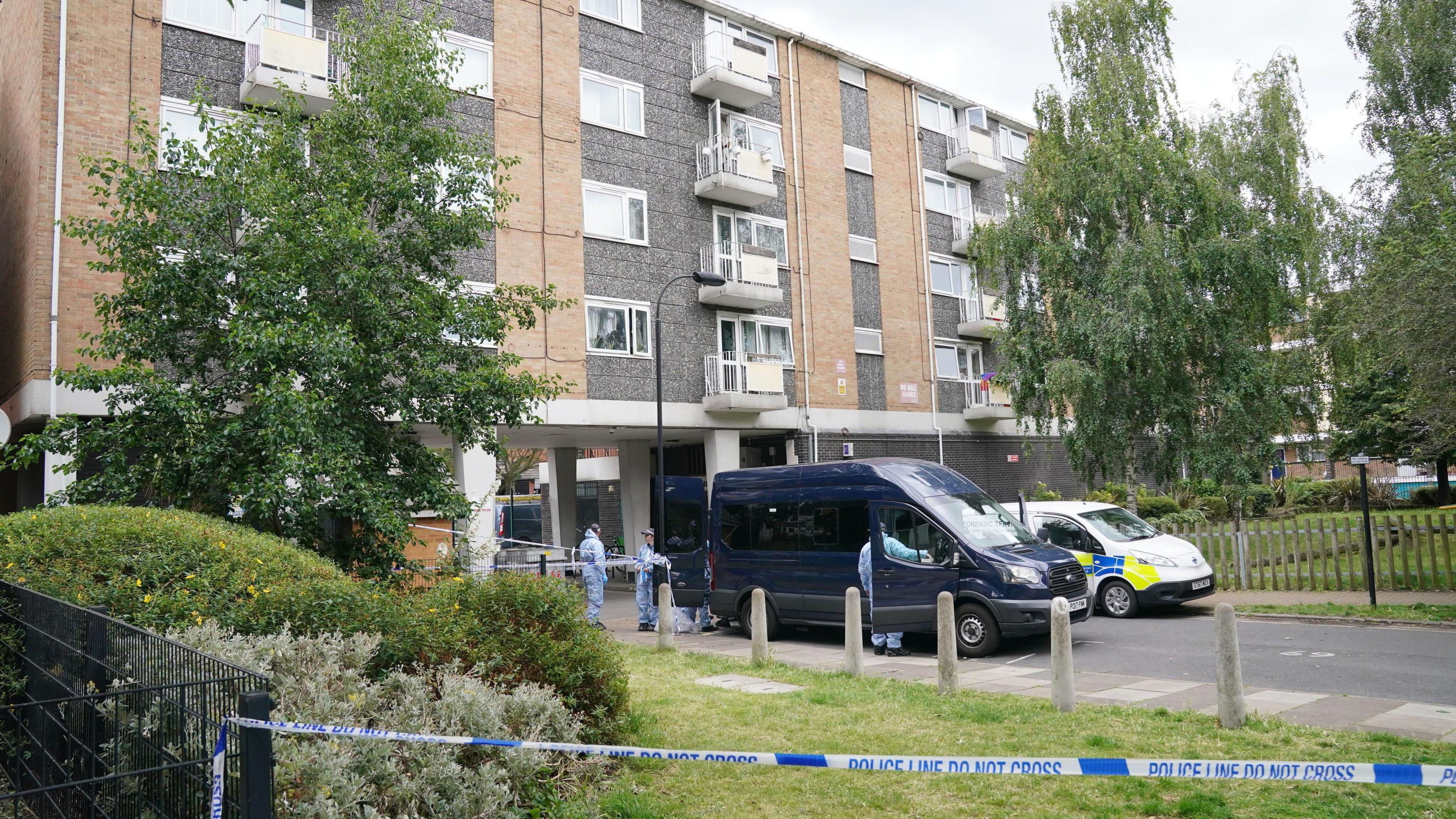 A police cordon in the foreground with forensic offices in blue overalls outside a block of flats