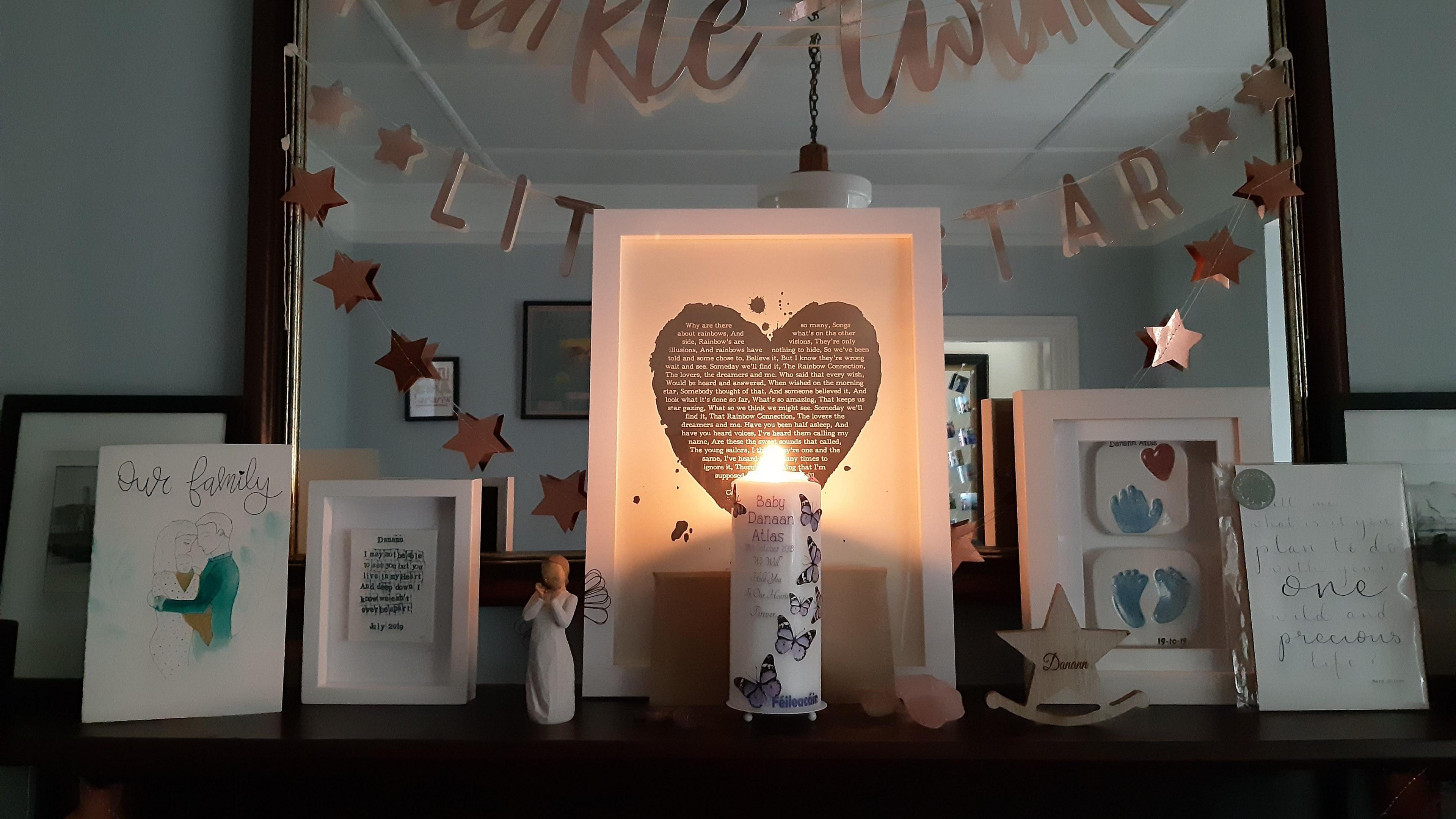 A photo of a mantlepiece in Maria Moynihan's home containing a lit candle decorated with her stillborn son Danann's name, alongside cards, a framed poem, a wooden star and framed casts of his hand and feet