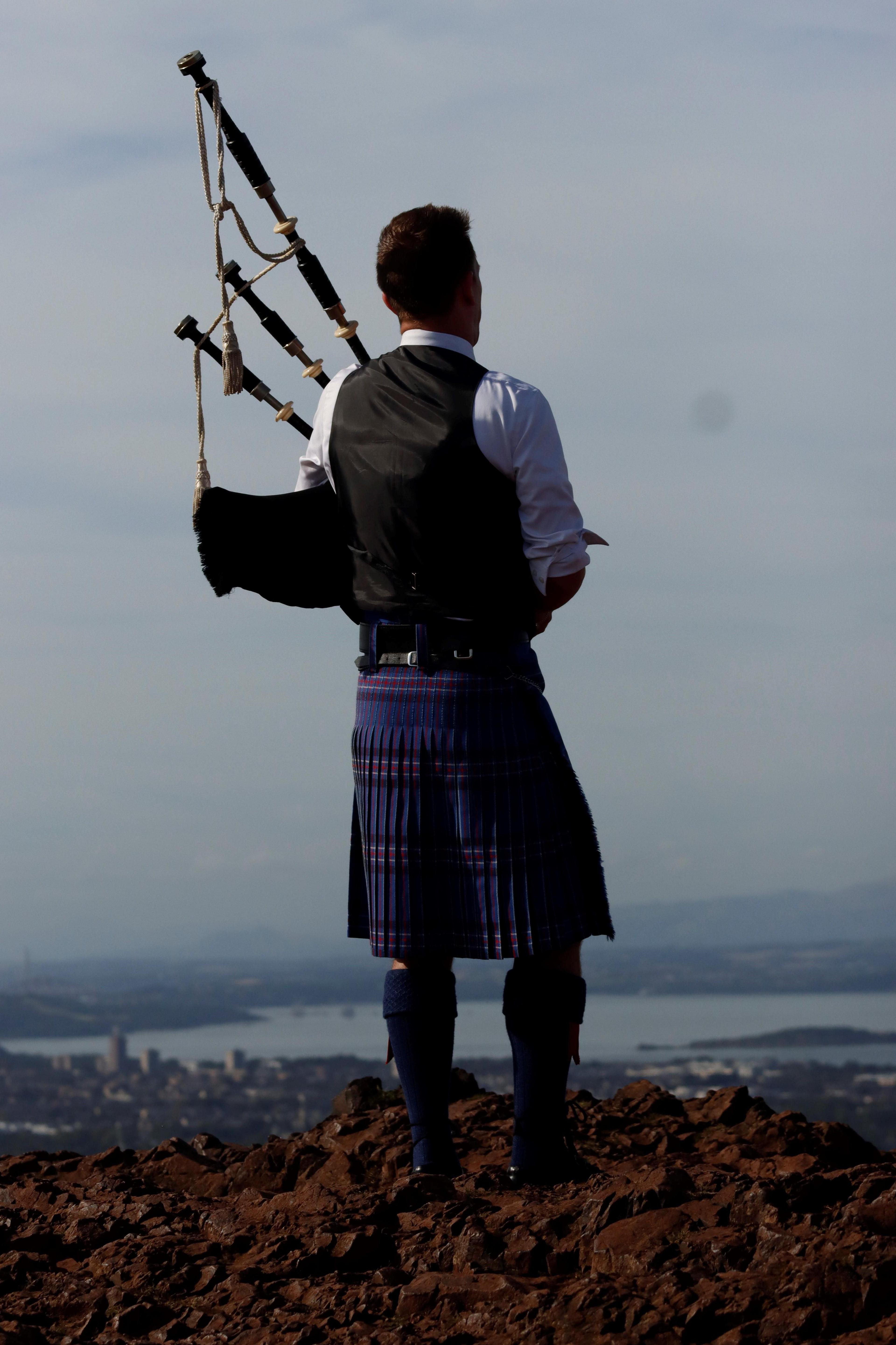 A man playing the bagpipes wearing a blue kilt and black waistcoat with a white shirt underneath. He is standing at the top of Arthur's Seat with a view of Edinburgh below.