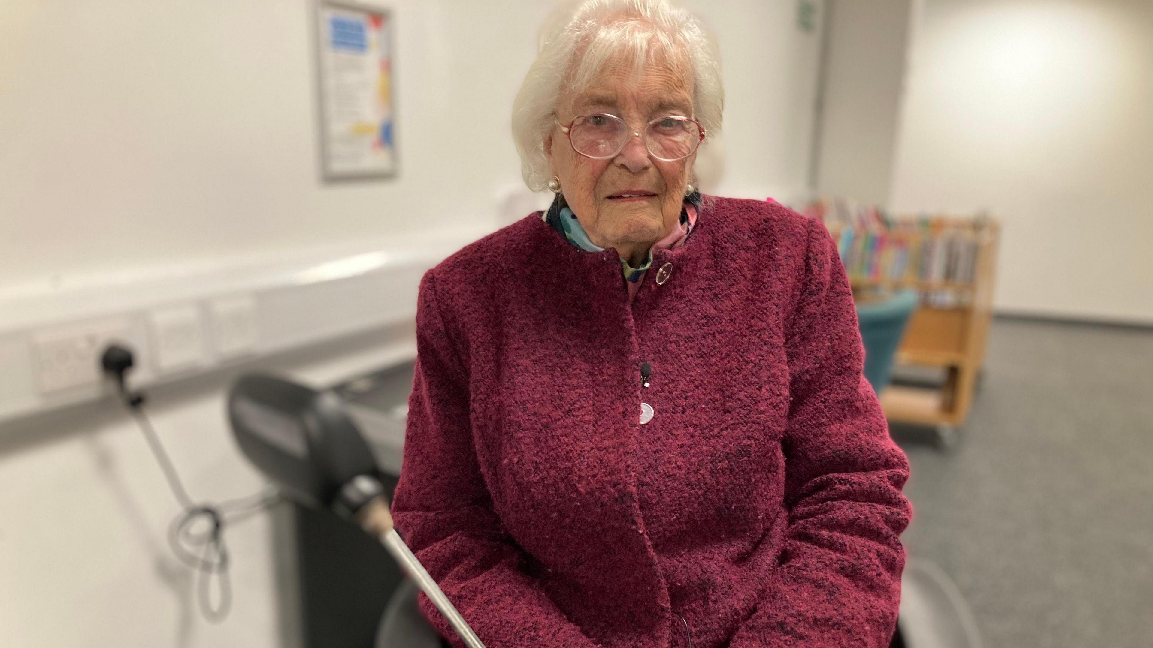 A pensioner with short grey hair and round glasses sits on a mobility scooter inside possibly a library room. She is wearing a dark red coat, there is a book trolley in the blurred background behind her.