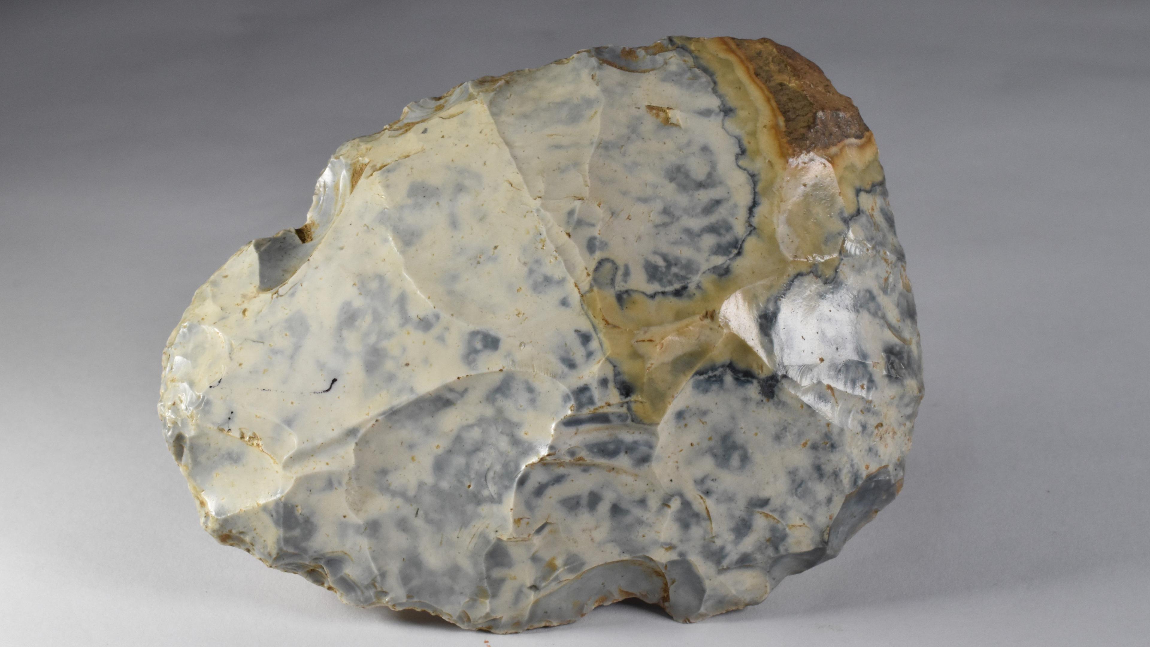 The handaxe rests on its side. It is a blue and white rock with some parts cut out of it.