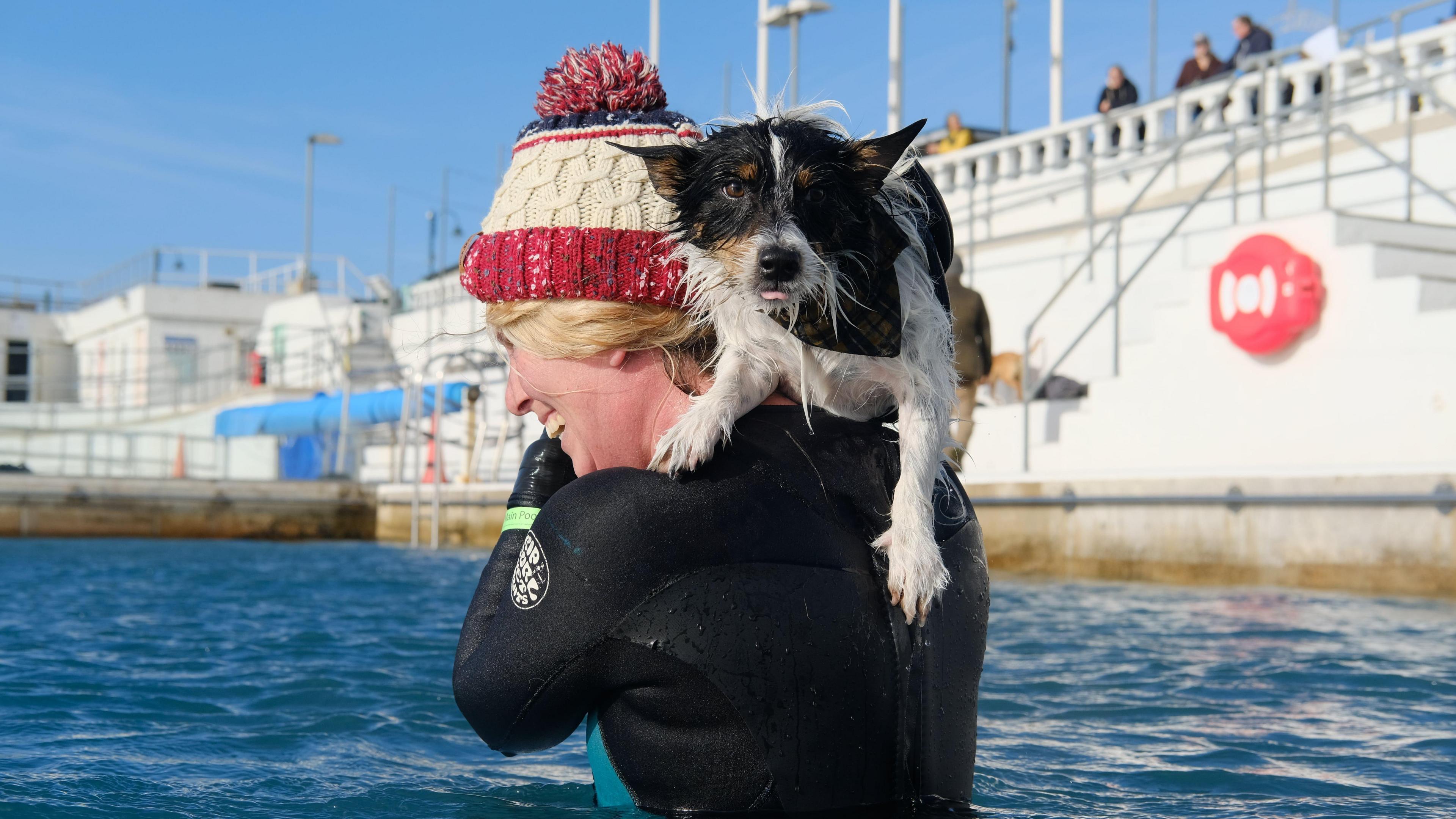 A black and white dog on the shoulder of a woman in a swimming pool. The woman is wearing a wetsuit and a red and white woolly hat.