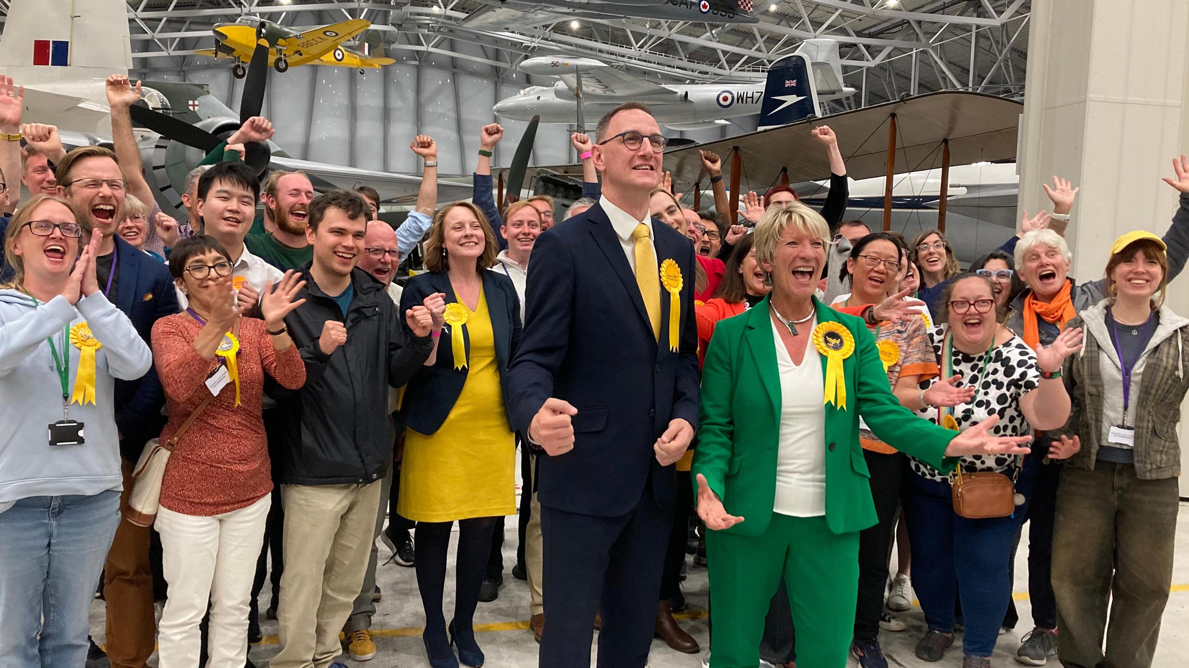 A crowd of people wearing Liberal Democrat rosettes cheer and celebrate.