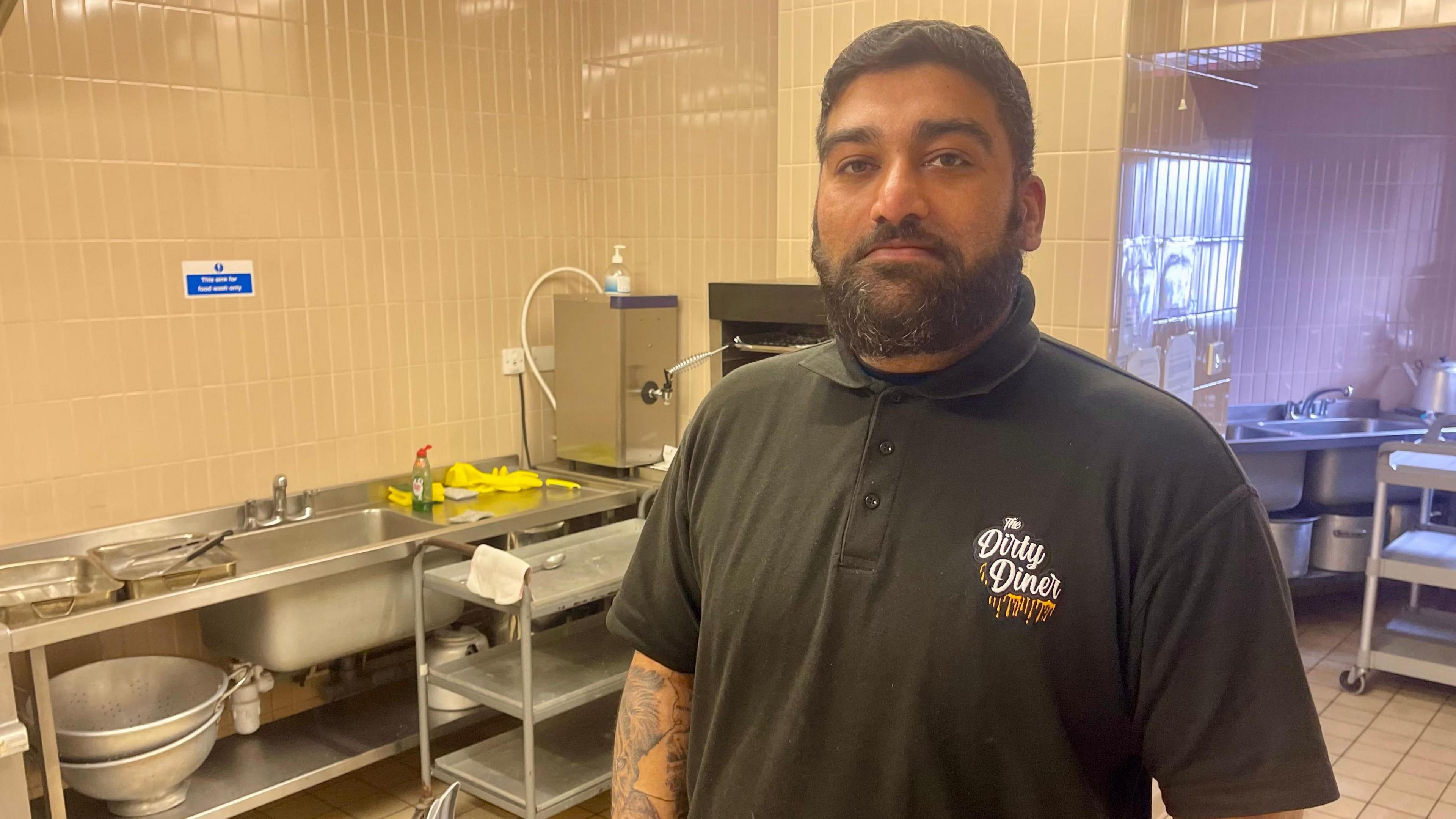 A man is stood in a tidy work kitchen. He has short dark hair and is wearing a black polo shirt with a Dirty Diner logo on the left. The kitchen is fitted out with cream tiles and there is a big stainless steel sink running along one wall.