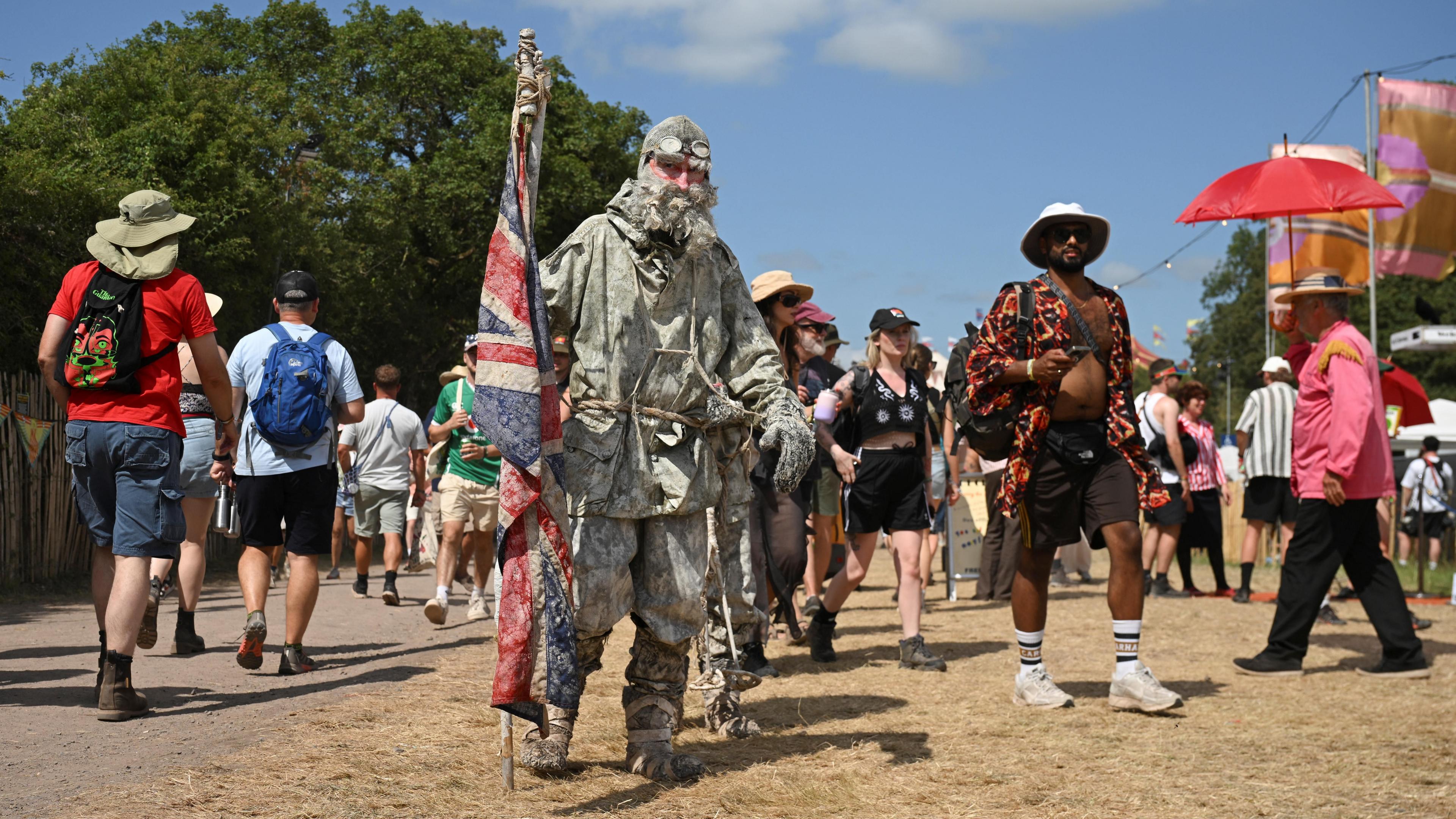 A person in the foreground is dressed in warm clothes, resembling an Arctic explorer. They are holding a British flag. Behind them, people are walking past in T-shirts and shorts wearing sun hats.