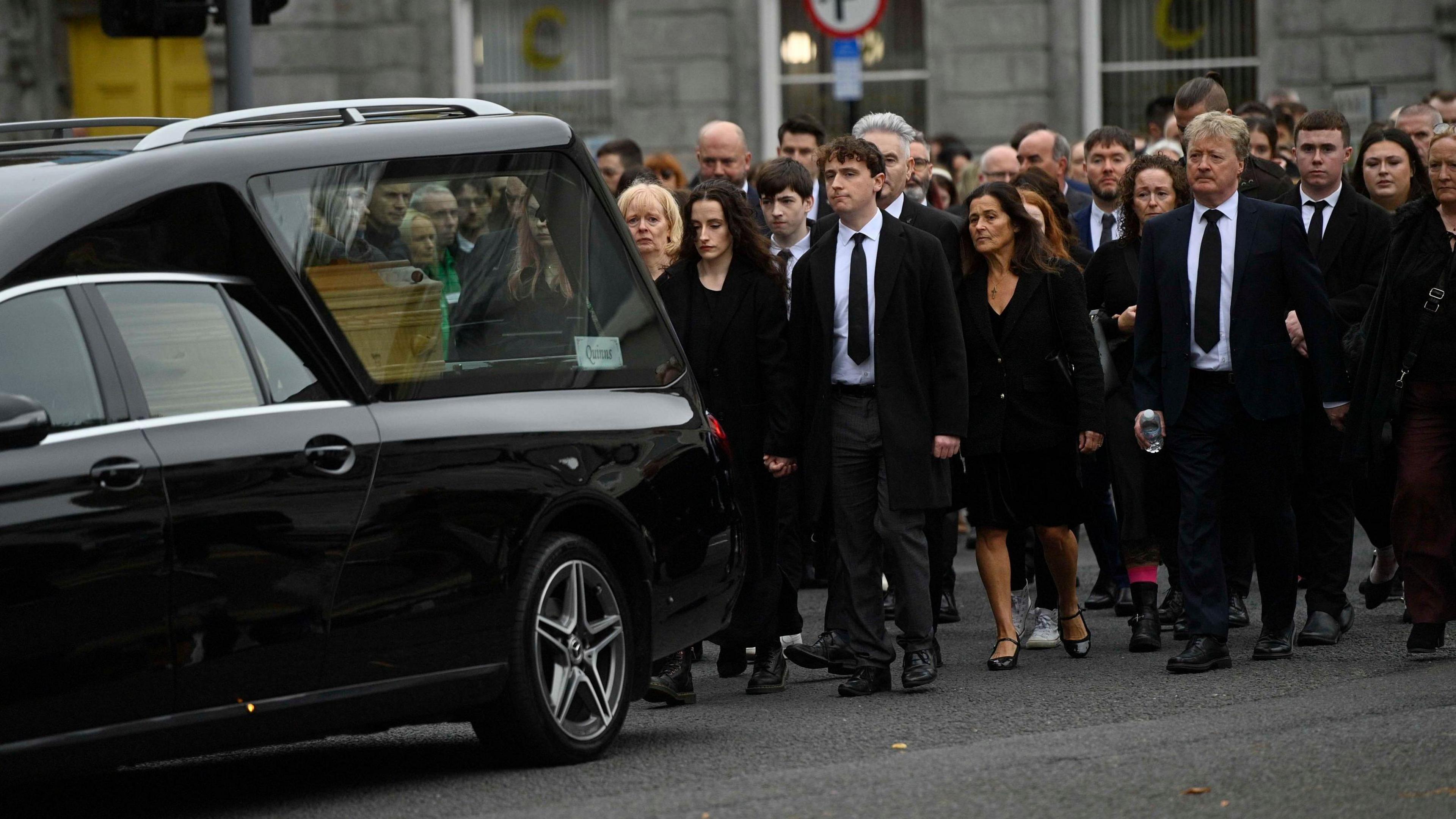 A hearse with a coffin inside is moving along a road with a procession of people, dressed in black, behind it.
