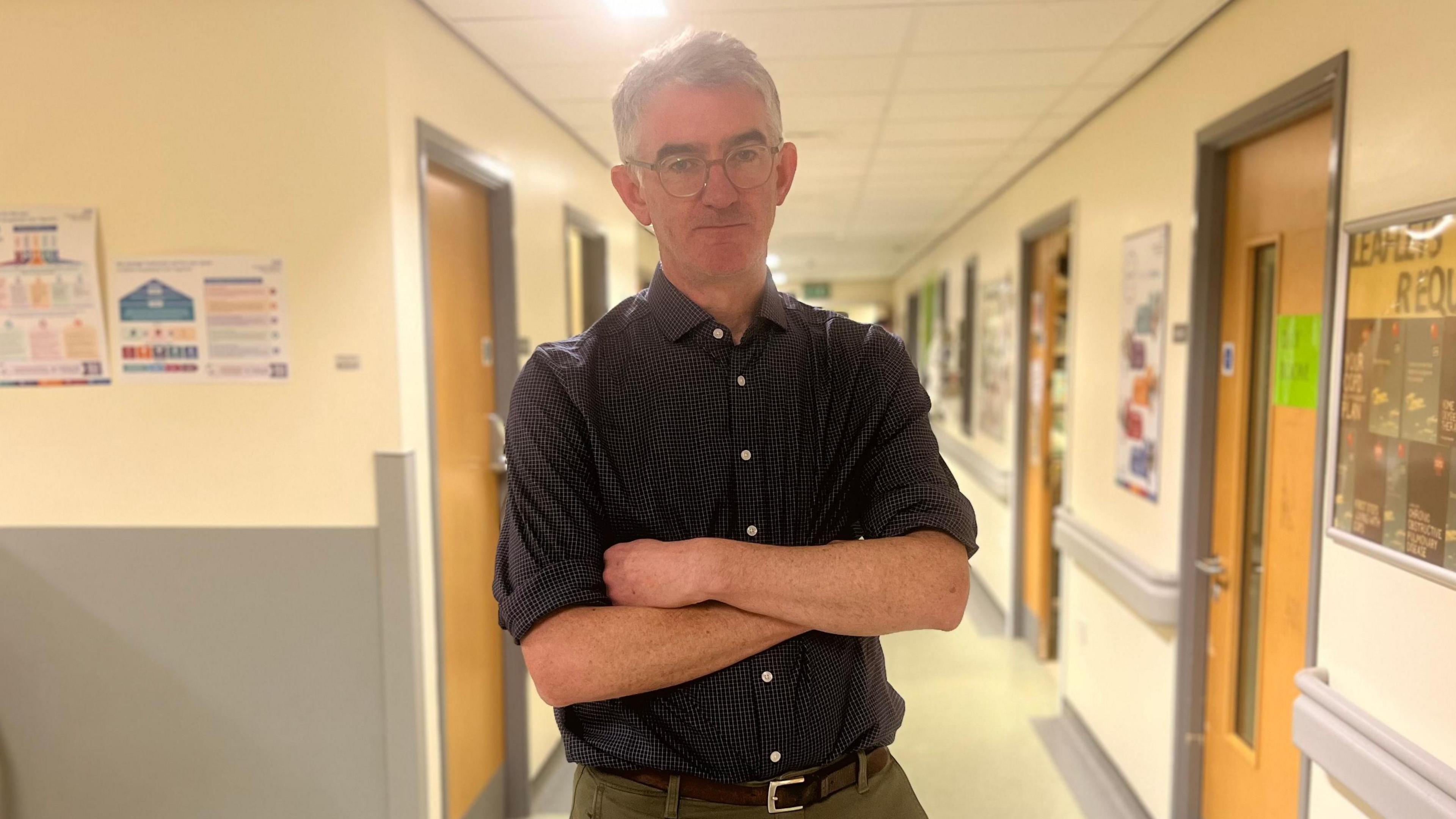 Man in blue shirt and glasses stands in a hospital ward corridor with his arms folded 