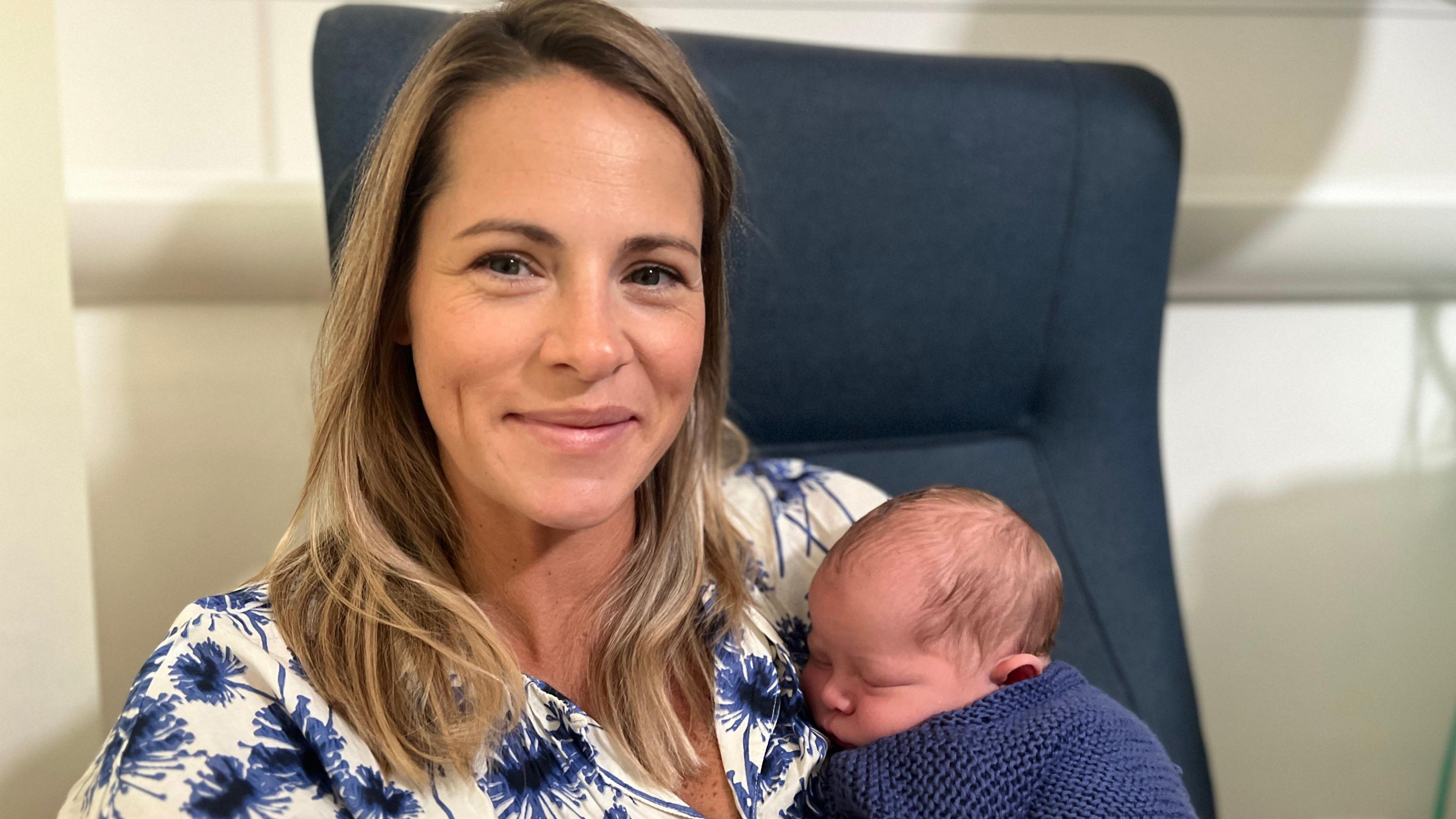 Jessica Williams with below shoulder-length fair hair, wearing a white top with large blue flowers, leaning against a blue chair. She is holding her son Orlando in her arms. He is sleeping and wrapped in a dark blue knitted jumper.