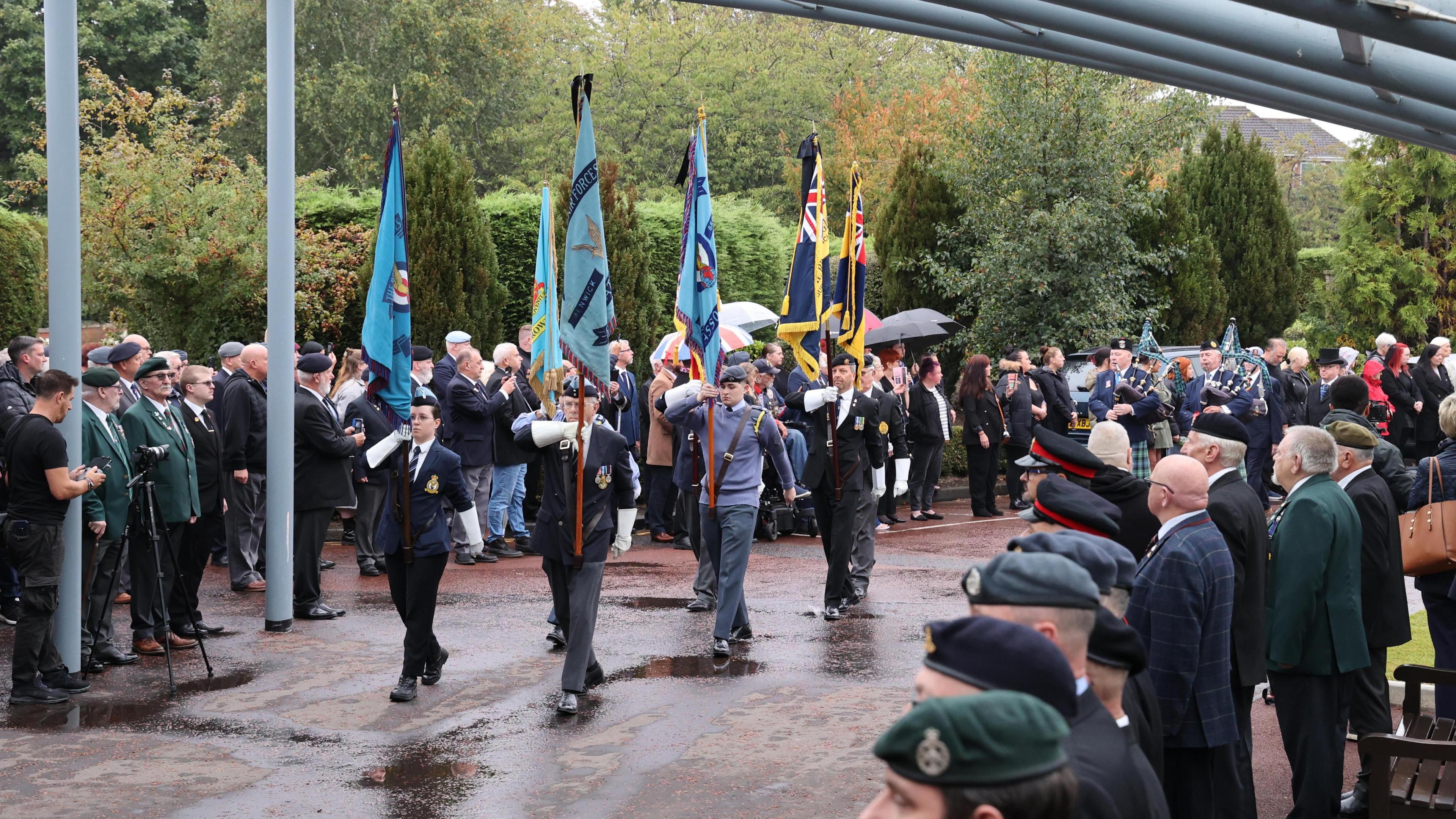 Half a dozen people representing the army, navy and RAF march towards the crematorium carrying flags as onlookers watch on. Some people in the crowd are holding umbrellas to shield them from rain.
