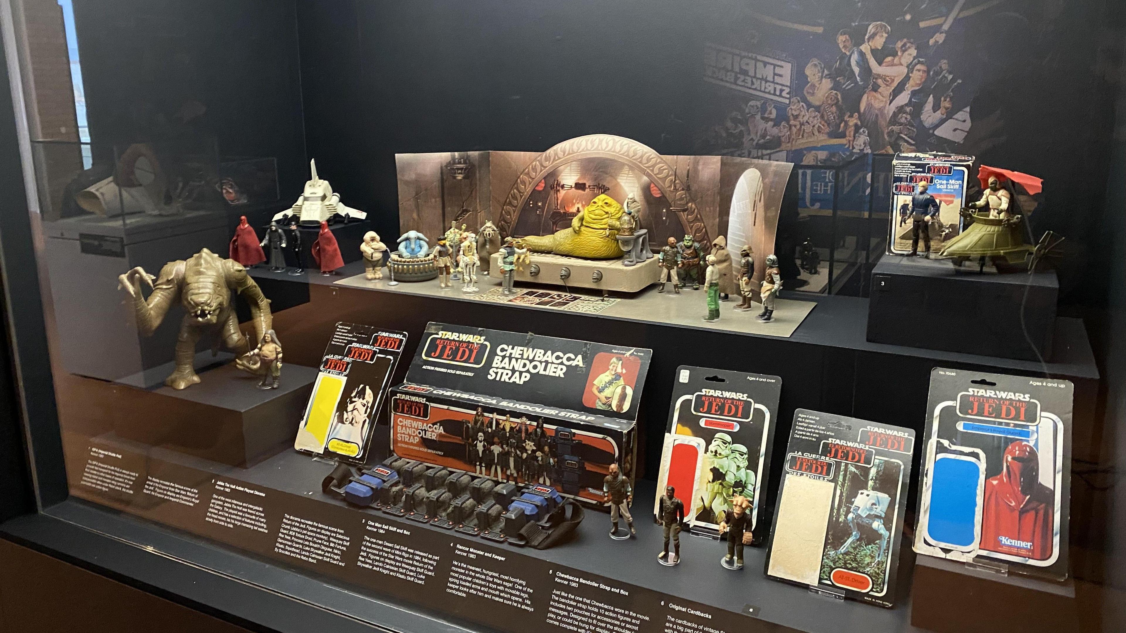 A museum display behind glass of vintage Star Wars toys from 1983's Return of the Jedi film. They include orange, slug-like crime lord Jabba the Hutt who is placed on a toy throne, a brown Rancor monster with claws outstretched, and an alien musical group known as The Max Rebo Band, which features a blue elephant-like creature sitting inside a circular piano-like instrument. There are various other action figures and small vehicles on display, including opened original packaging which once contained such figures.