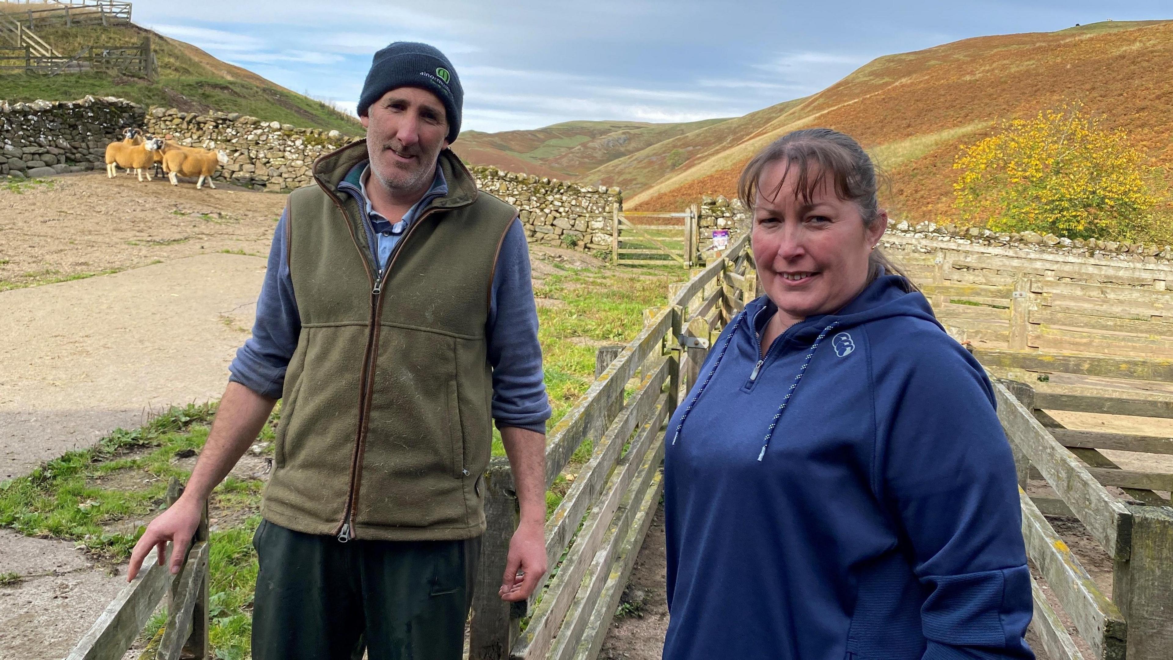 The image shows Dan and Sam Wood from Shillmoor in Northumberland standing in a farmyard with sheep in the rear of the shot and rolling hills behind 