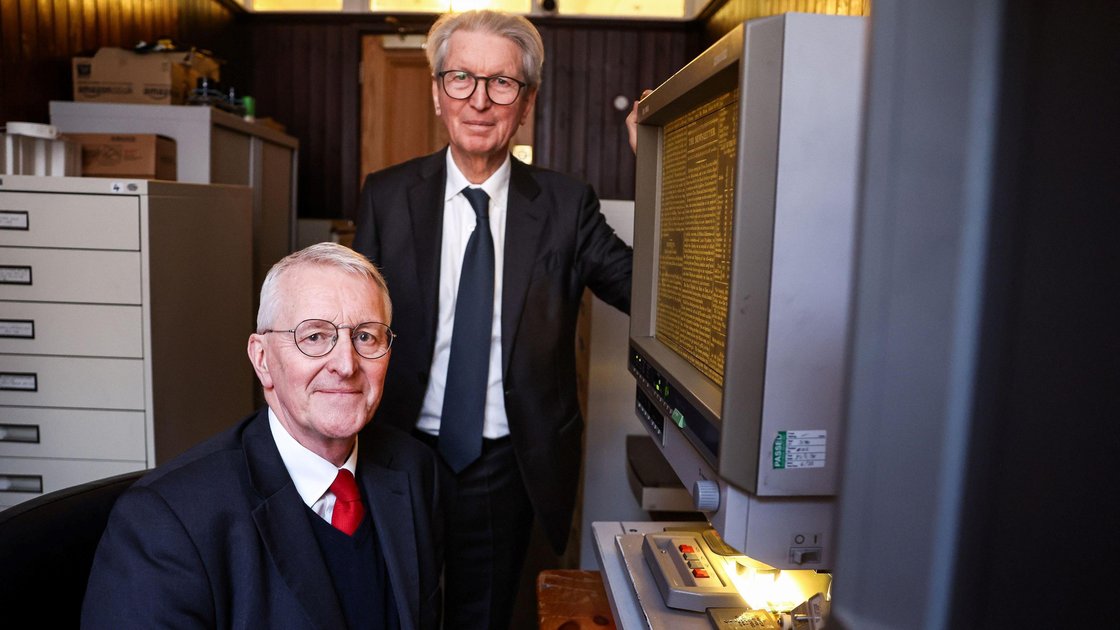 A man wearing glasses and a suit is sitting looking at the camera. Another man, also wearing glasses and a suit, is standing next to him. A large grey computer with archived copy is next to them. Filing cabinets are in the background. 