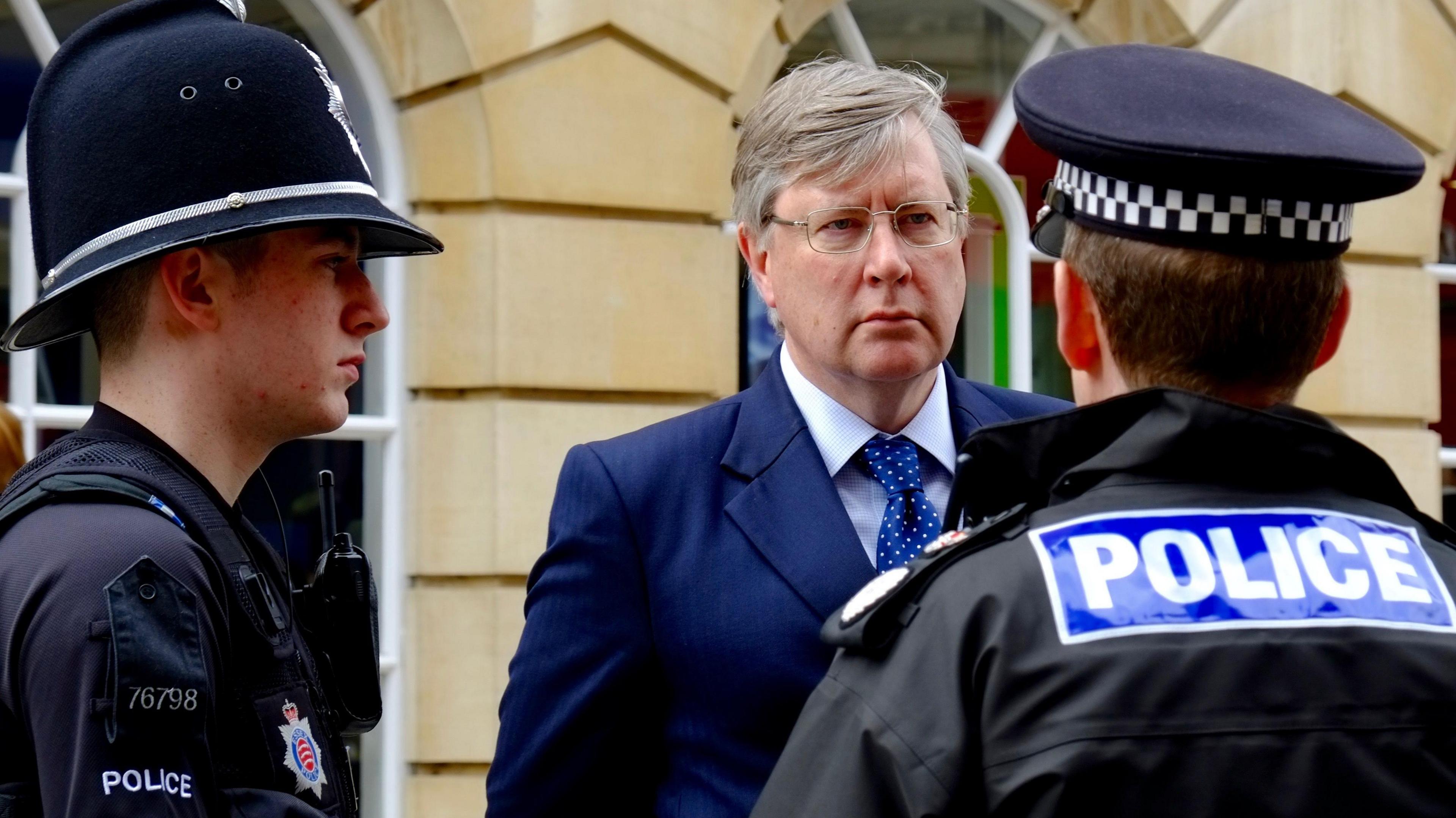 Roger Hirst is wearing a suit and a blue Conservative rosette while standing in a sports hall. 