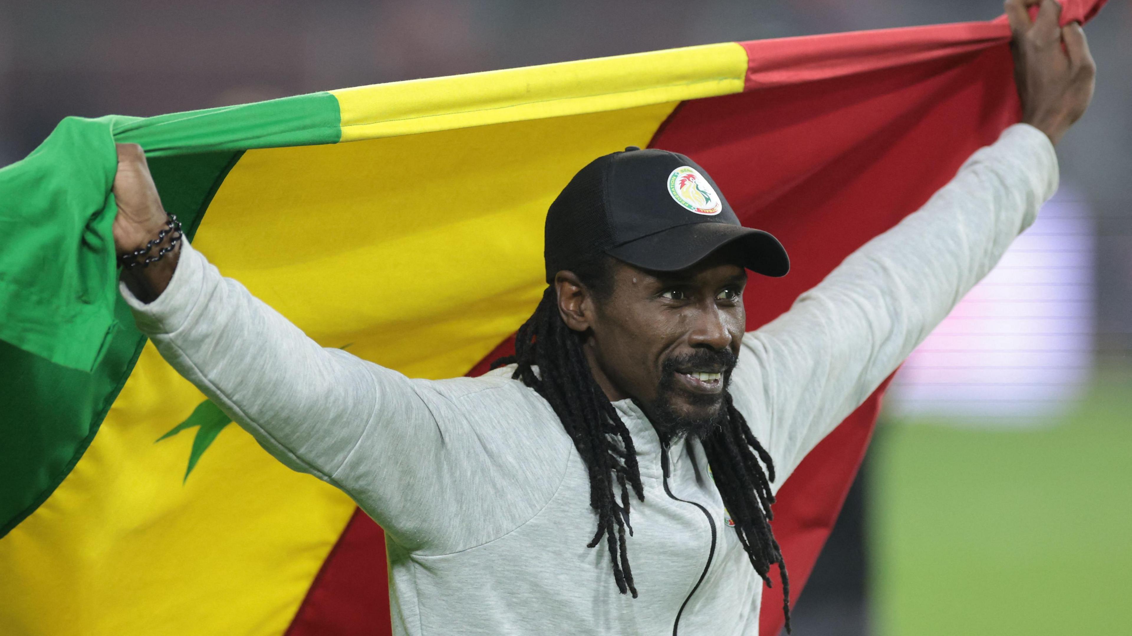 Aliou Cisse wearing grey tracksuit top and black cap with Senegal crest on it holds aloft a Senegal flag behind him as he moves across a football pitch