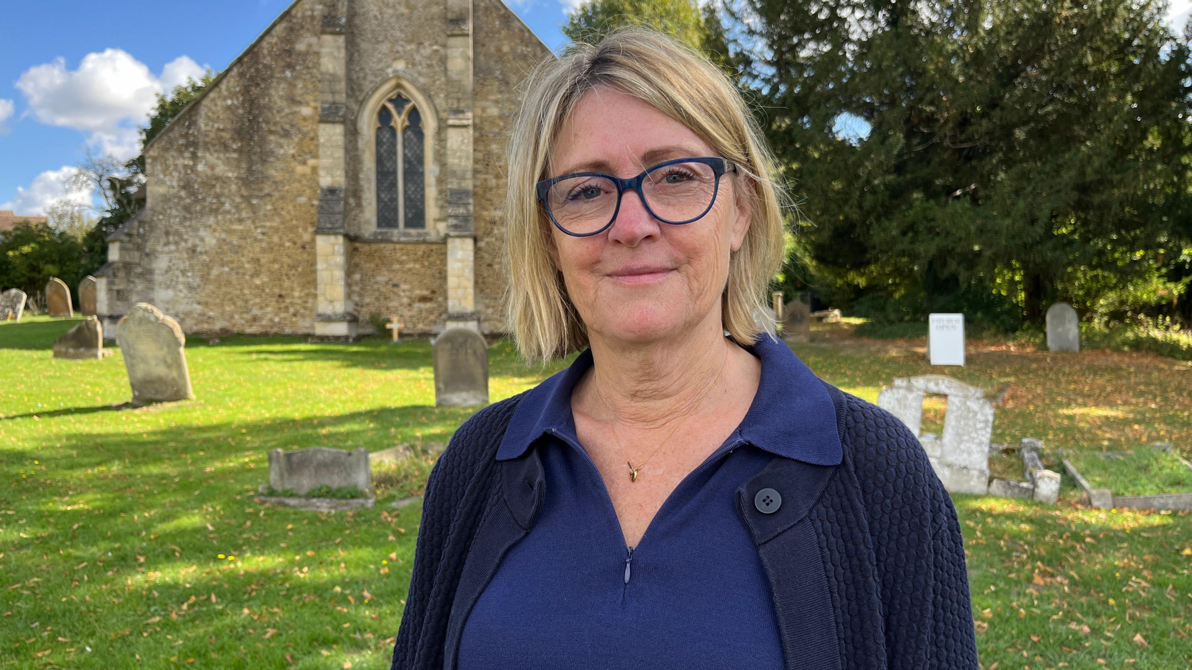 Narelle Hassell is standing in front of St Michael's Church in Longstanton. The church is made of stone and has a stained glass window. There are old gravestones on the grass. Ms Hassell has bob-length blond hair. She is wearing blue-rimmed spectacles and a blue top and cardigan