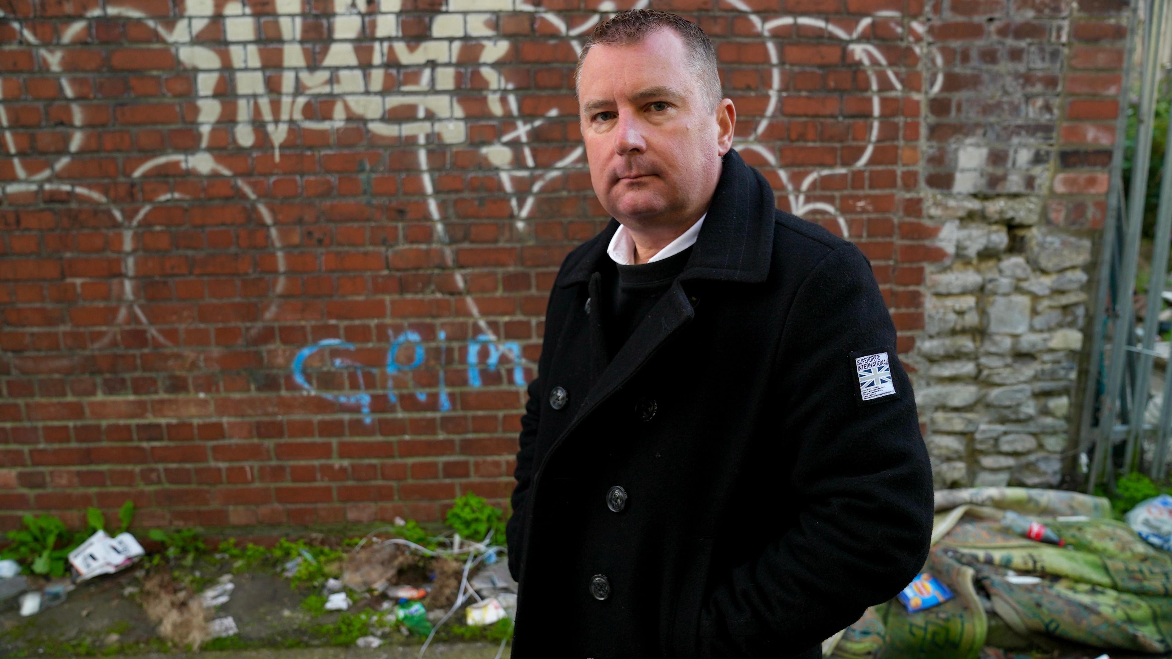 A man looking at the camera. He has a black coat on and there is a wall covered in graffiti behind him.