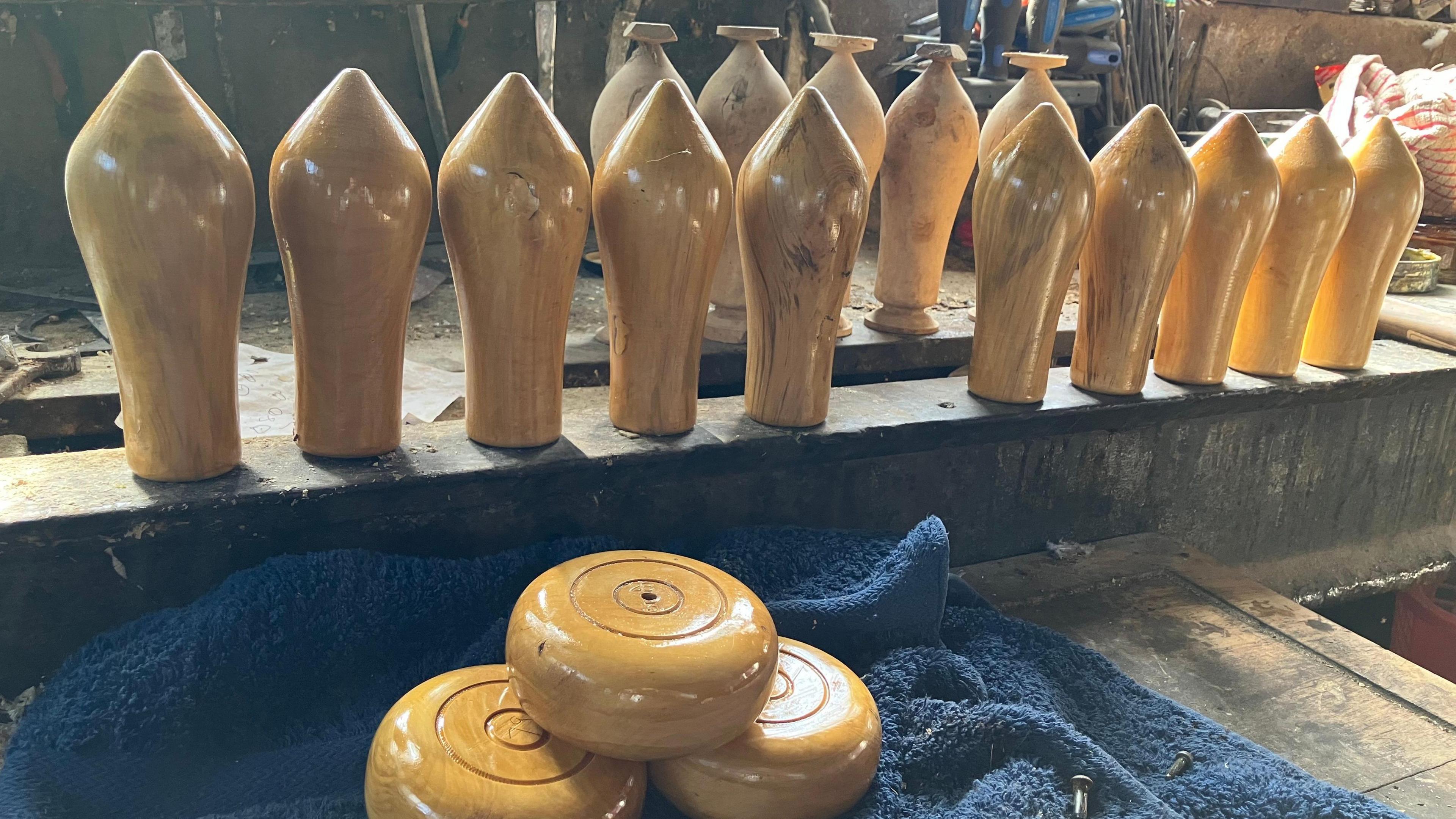 Ten varnished skittles stand on a platform in a workshop. Three wooden cheeses sit on a blue towel.