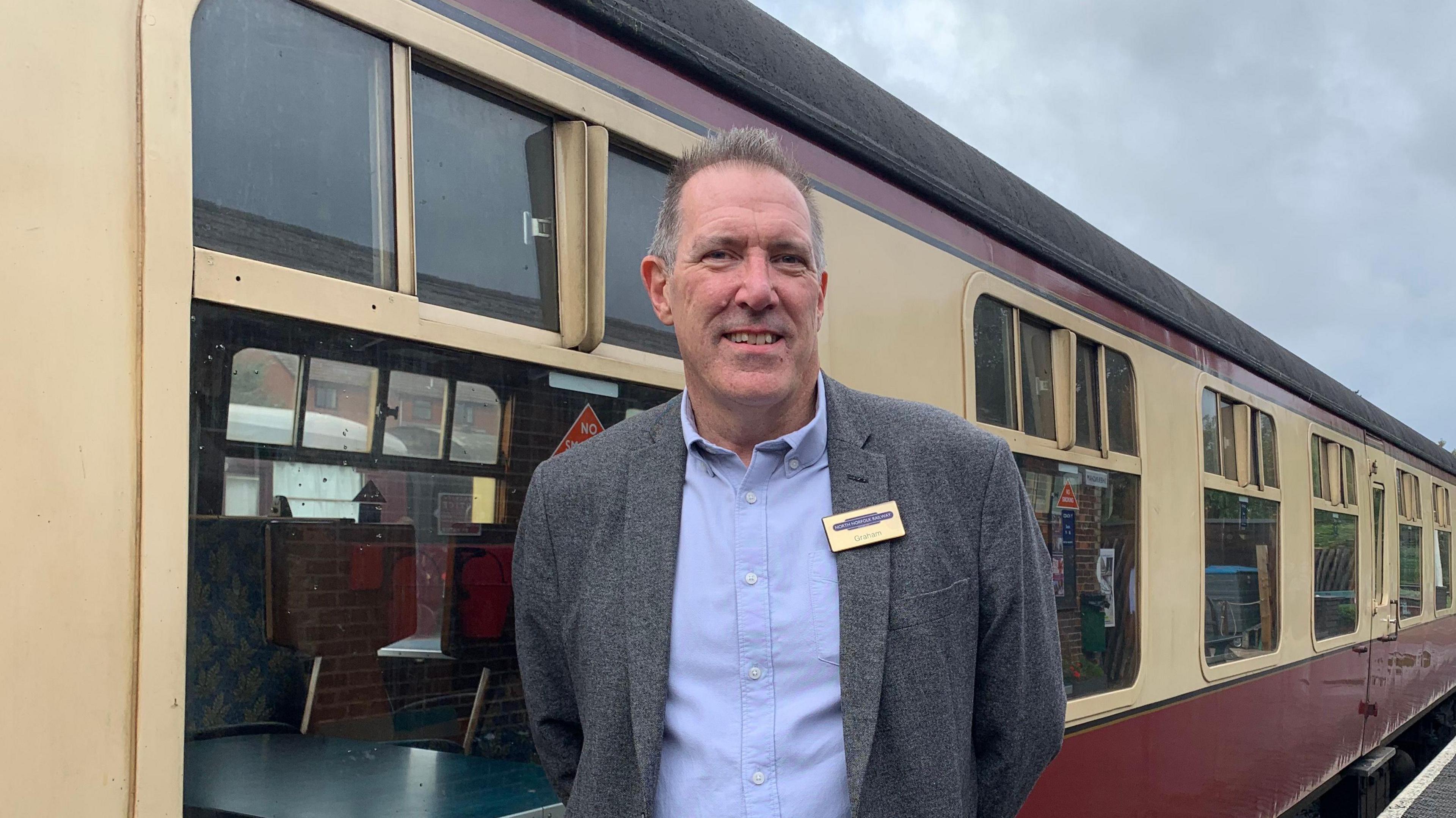 A man wearing a grey blazer and light blue shirt wearing a name badge is pictured stood in front of a yellow and red train carriage at a railway platform.