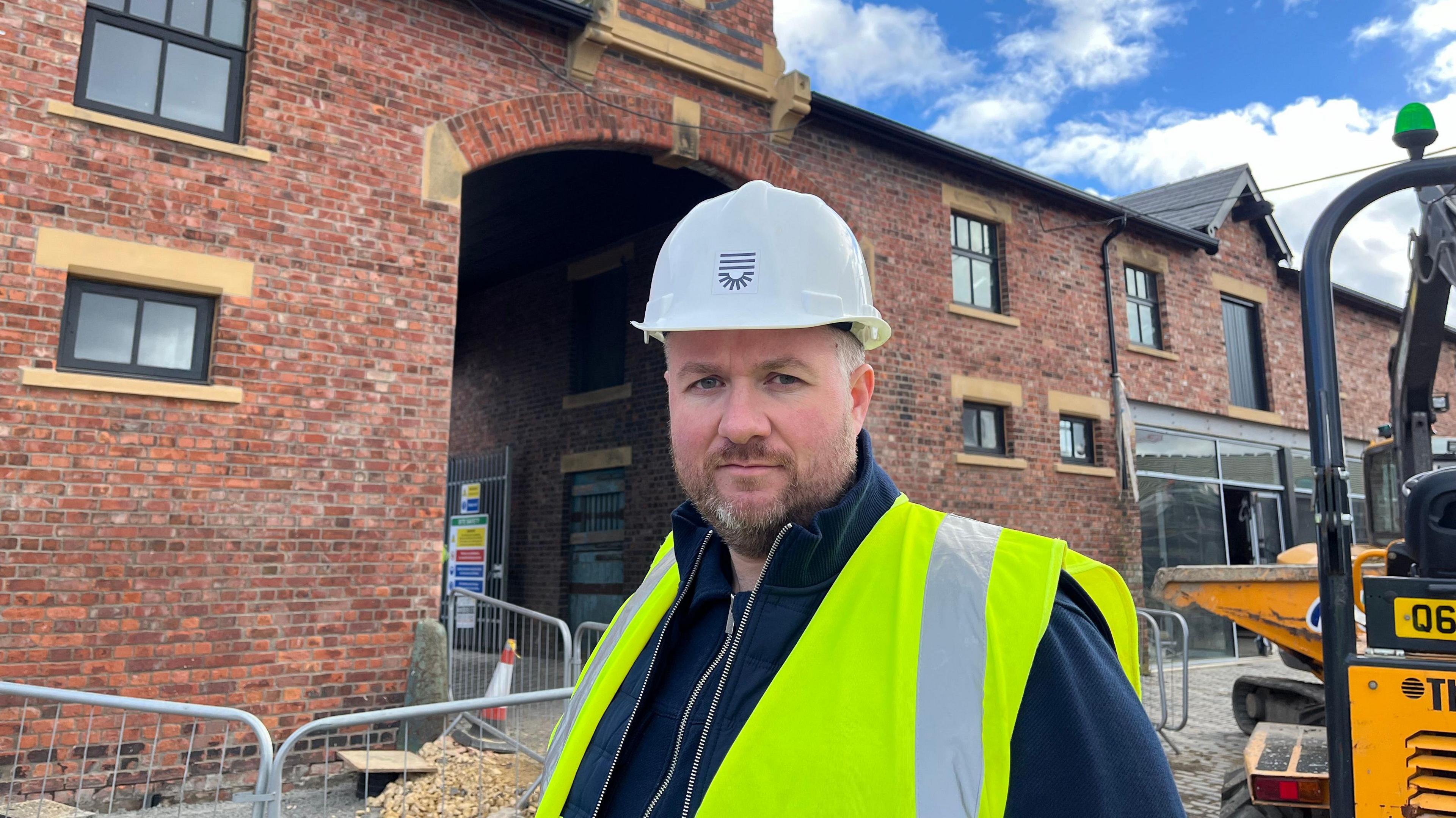 A man in a high vis jacket standing in front of a building with a digger to one side 