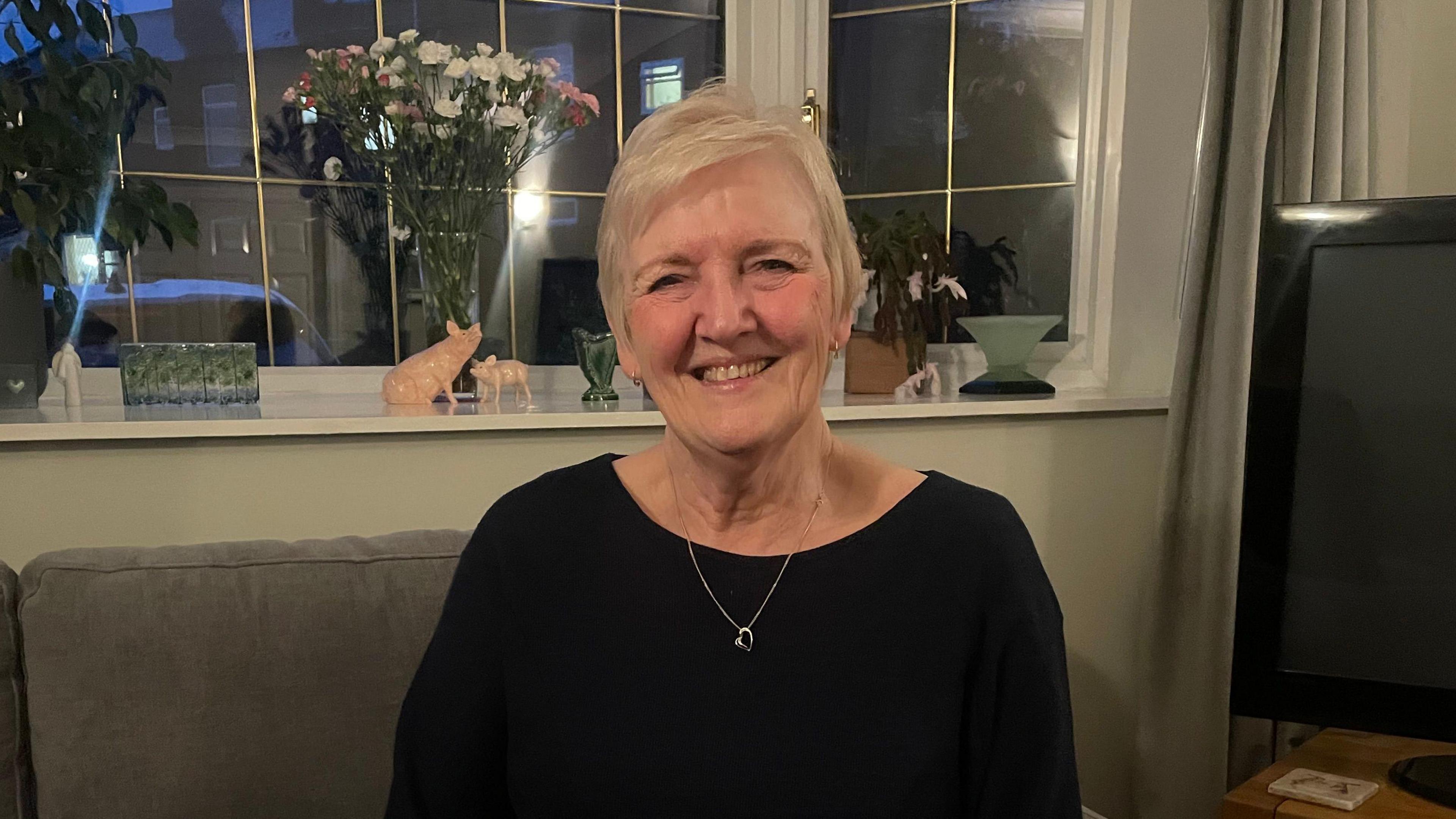 A smiling woman with short white-blonde hair sitting in a front room. She is smiling and wearing a silver necklace and black top. Behind her are some porcelain pig statues and a vase of white and pink flowers.
