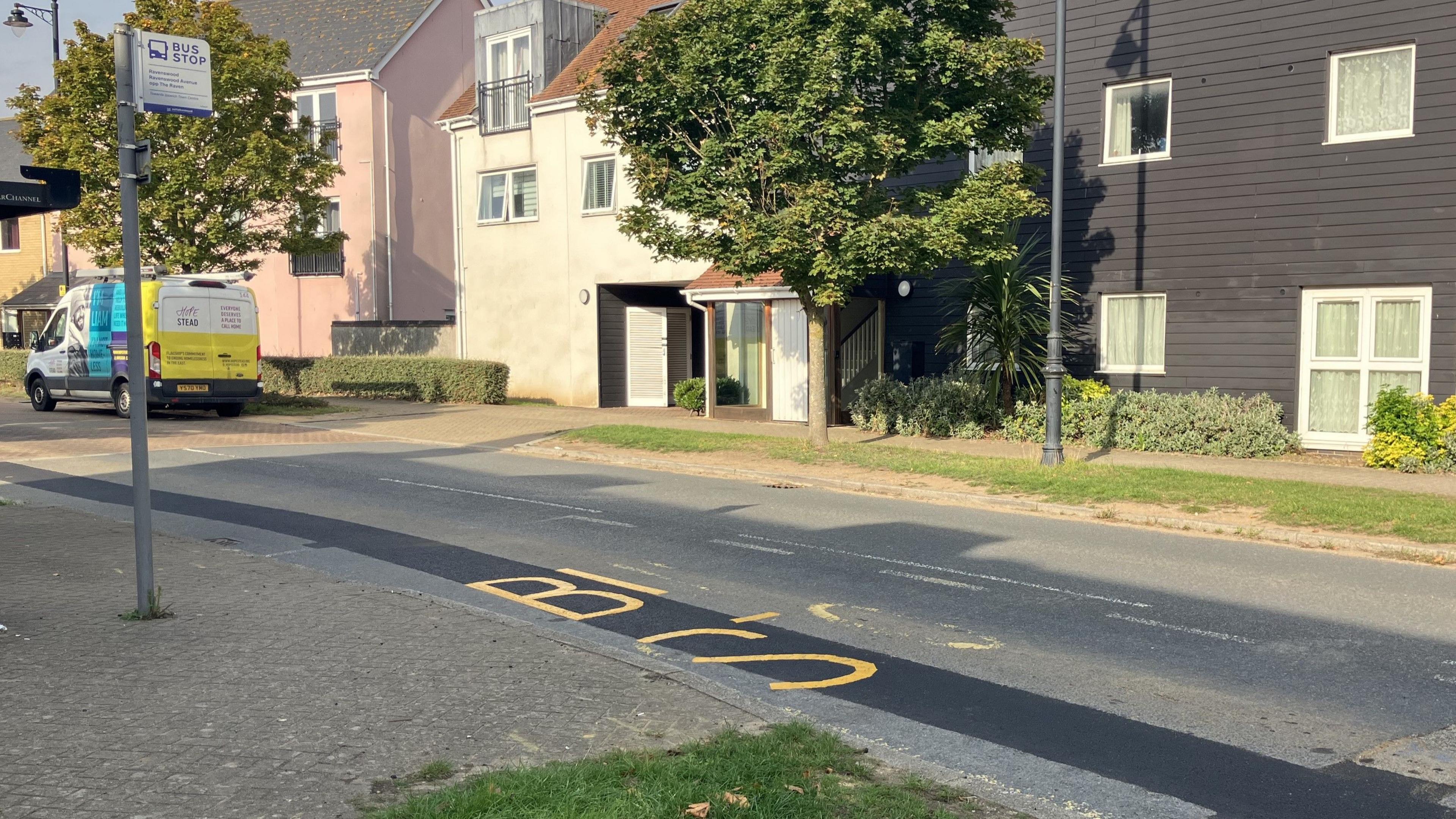 A wider view of the bus stop road marking that has only been partially repainted over. A bus stop can be seen on the pavement and on the other side of the road there are houses and flats. A van is parked on the other side of the road. 