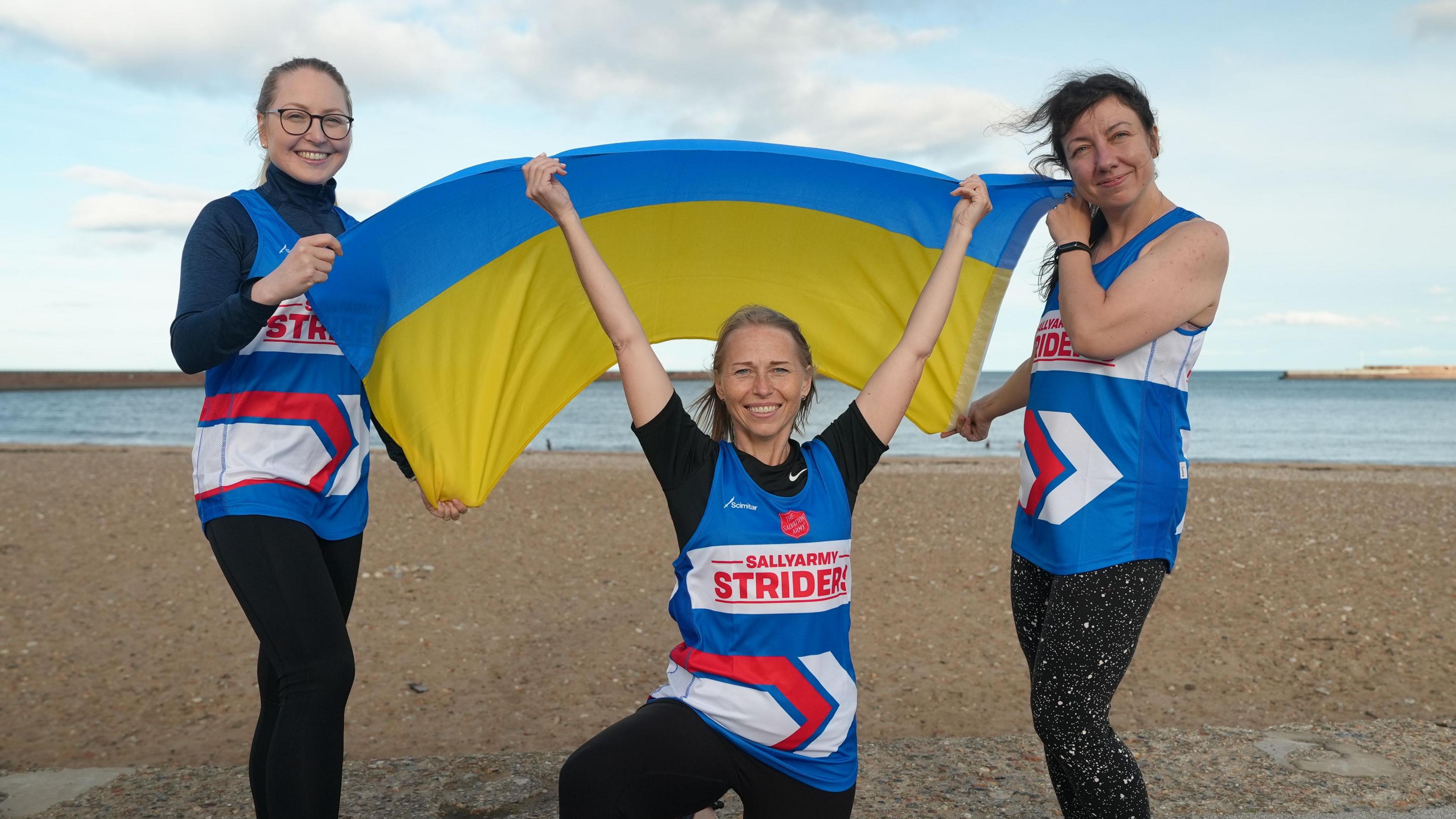 Three women dressed in running vests and leggings are smiling at the camera, they are holding a Ukrainian flag. The photograph is taken during training at the beach in Sunderland, the sand and sea can be seen in the background of the image.