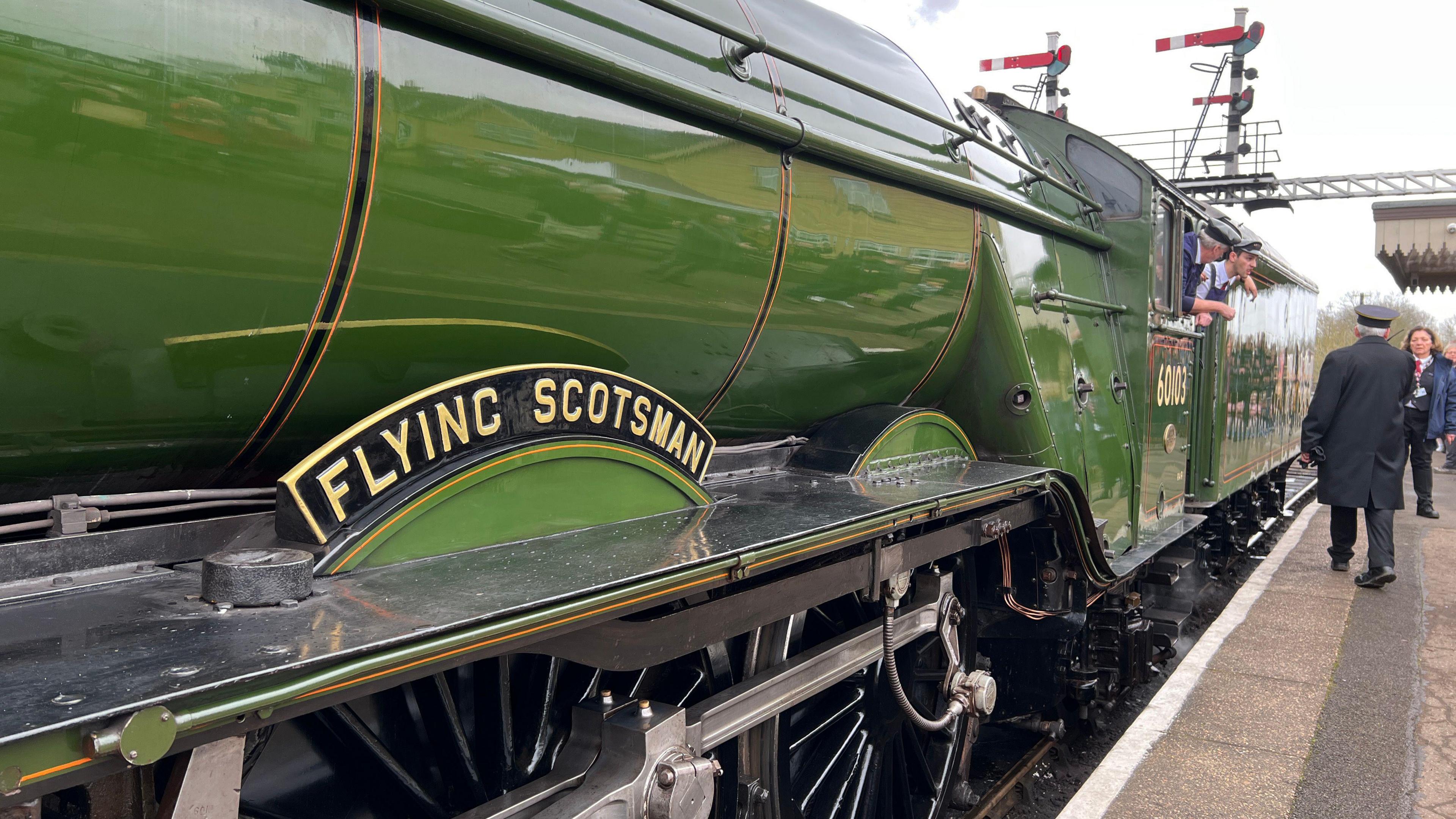 A shot of the Flying Scotsman from the side. It is painted green colour and has the words "Flying Scotsman" written on it. A conductor is walking up the platform next to it as two mean hang out of an open window and doorway.