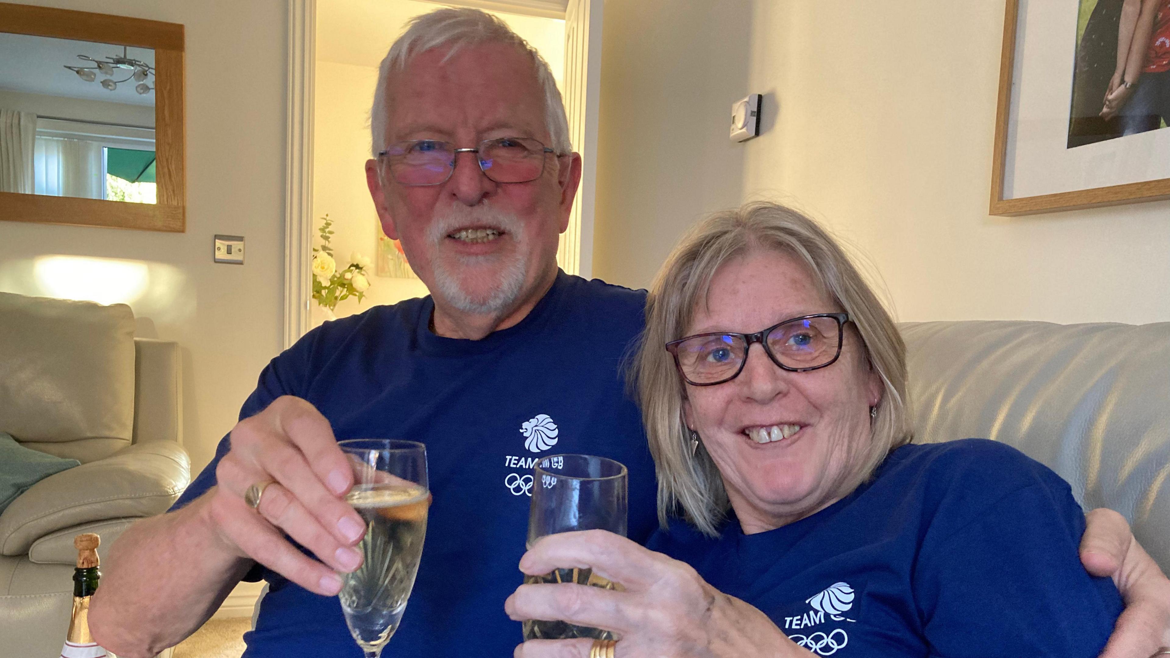 A man and a woman, both wearing navy blue Team GB tshirts, hold up glasses of Champagne while smiling