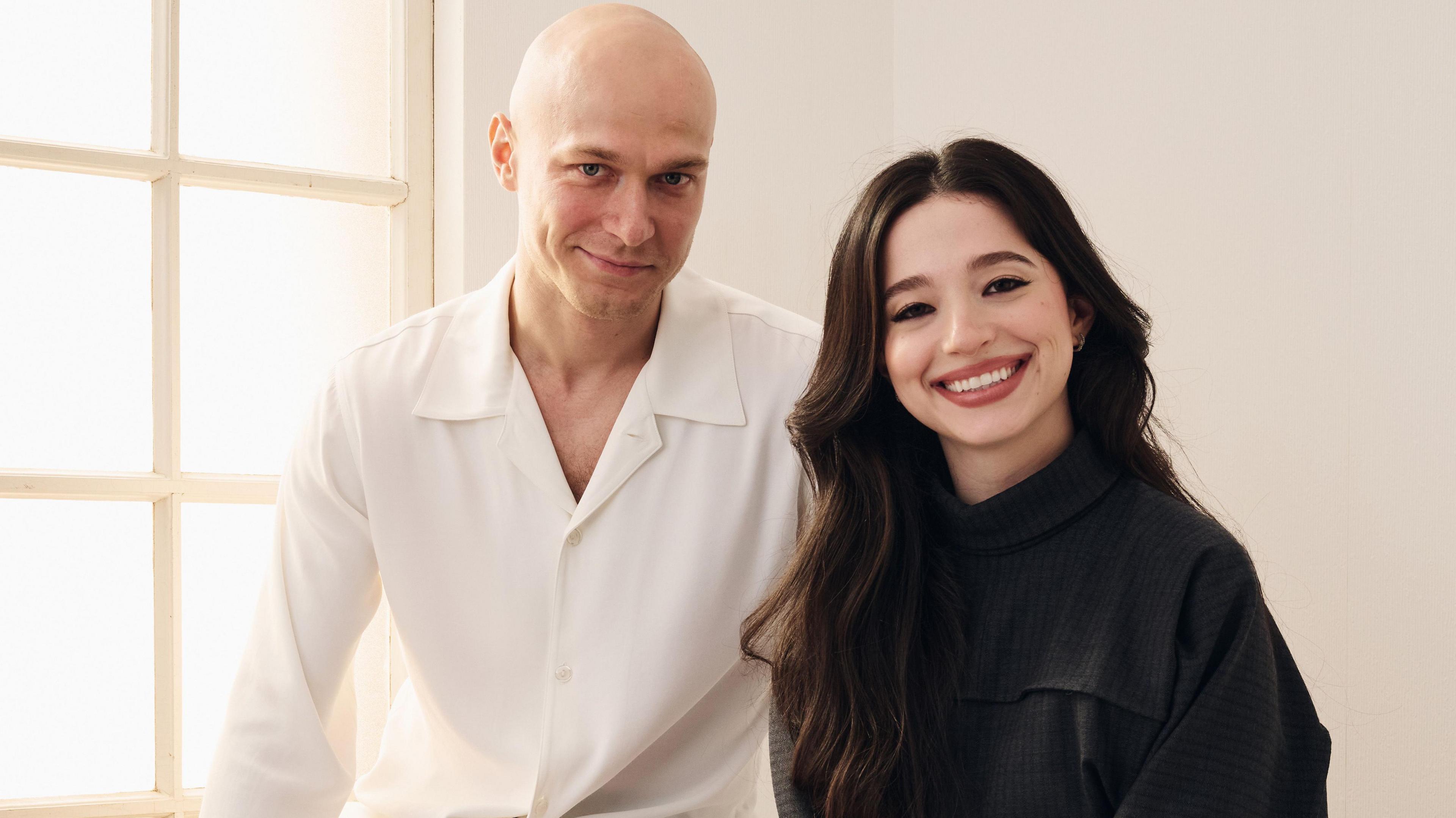 Yura Borisov and Mikey Madison of Anora at the Toronto International Film Festival. They are standing in front of a window. Borisov is wearing a white shirt, while Madison is dressed in black.