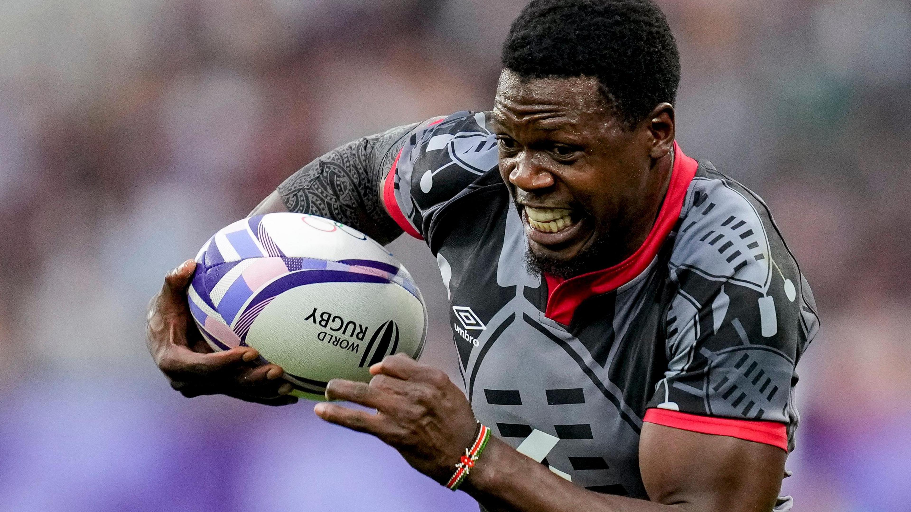 Vincent Onyala, wearing a grey Kenya shirt with red trim, grimaces as he runs forward holding a rugby ball in his right hand