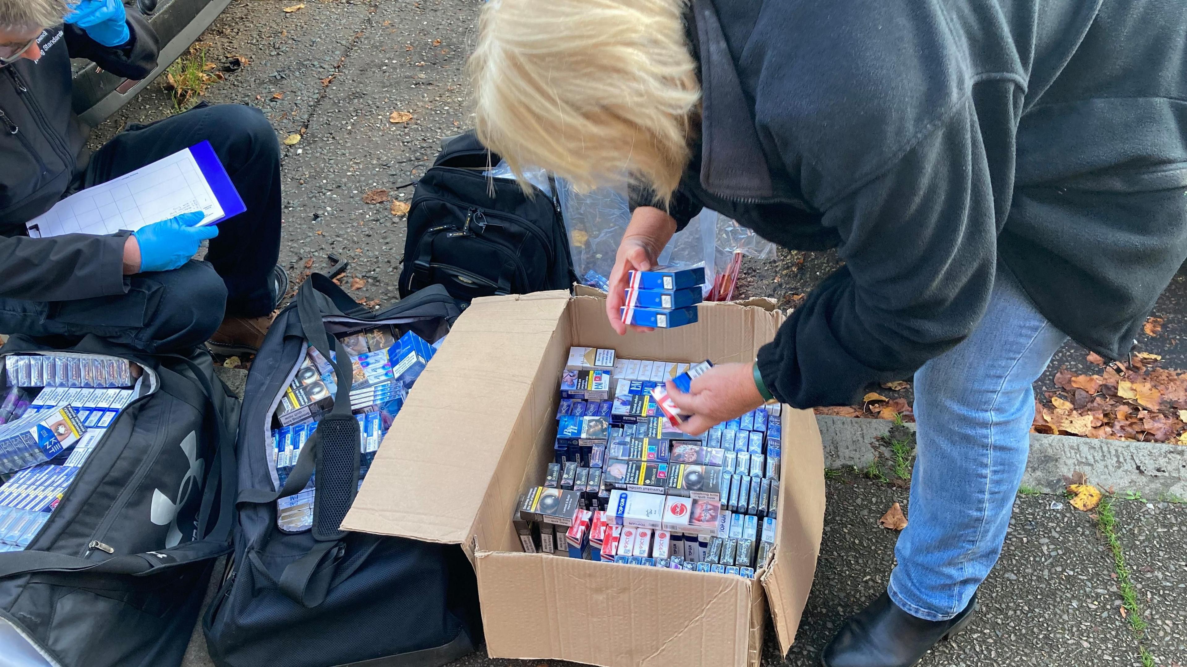 Boxes of illegal cigarettes and tobacco in a box and two black holdalls on the pavement