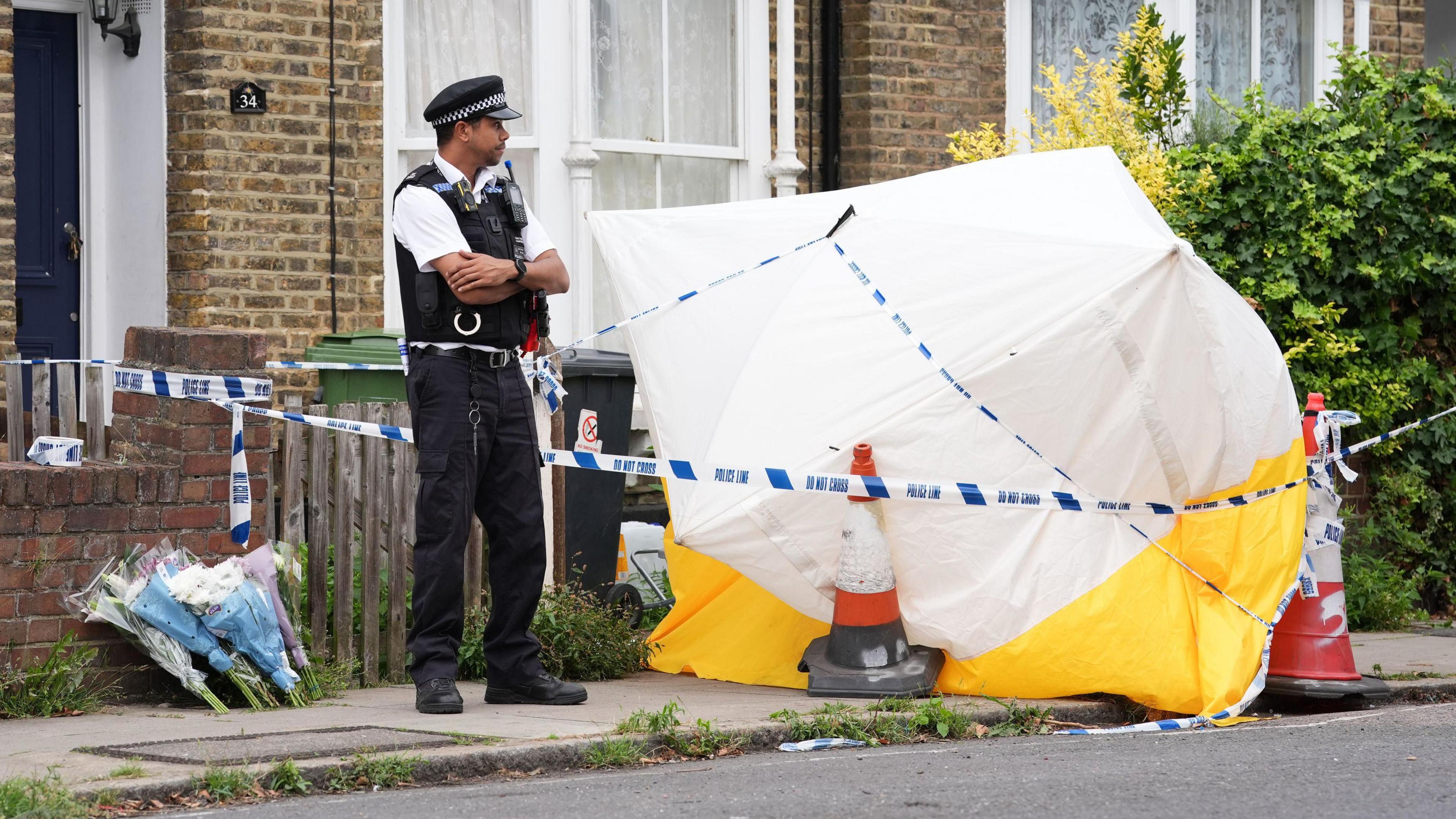 A police officer stands in front of a police cordon and a white forensic tent outside a house on Monson Road in New Cross. A few bunches of flowers have been left on the pavement.