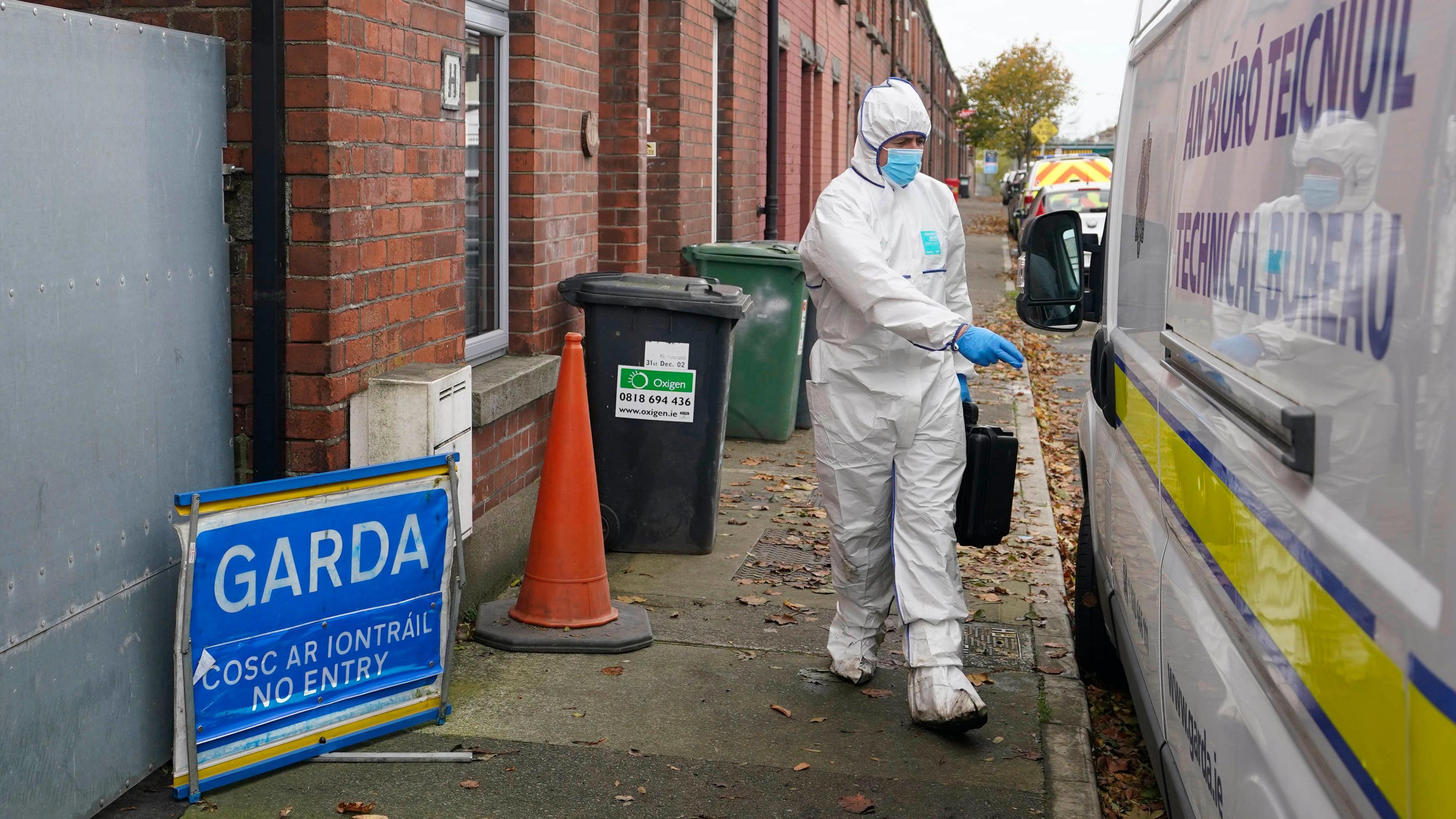 A Garda forensic officer, wearing white overalls, walking along a path outside red brick houses. There is a white Garda van to the left of the image. 