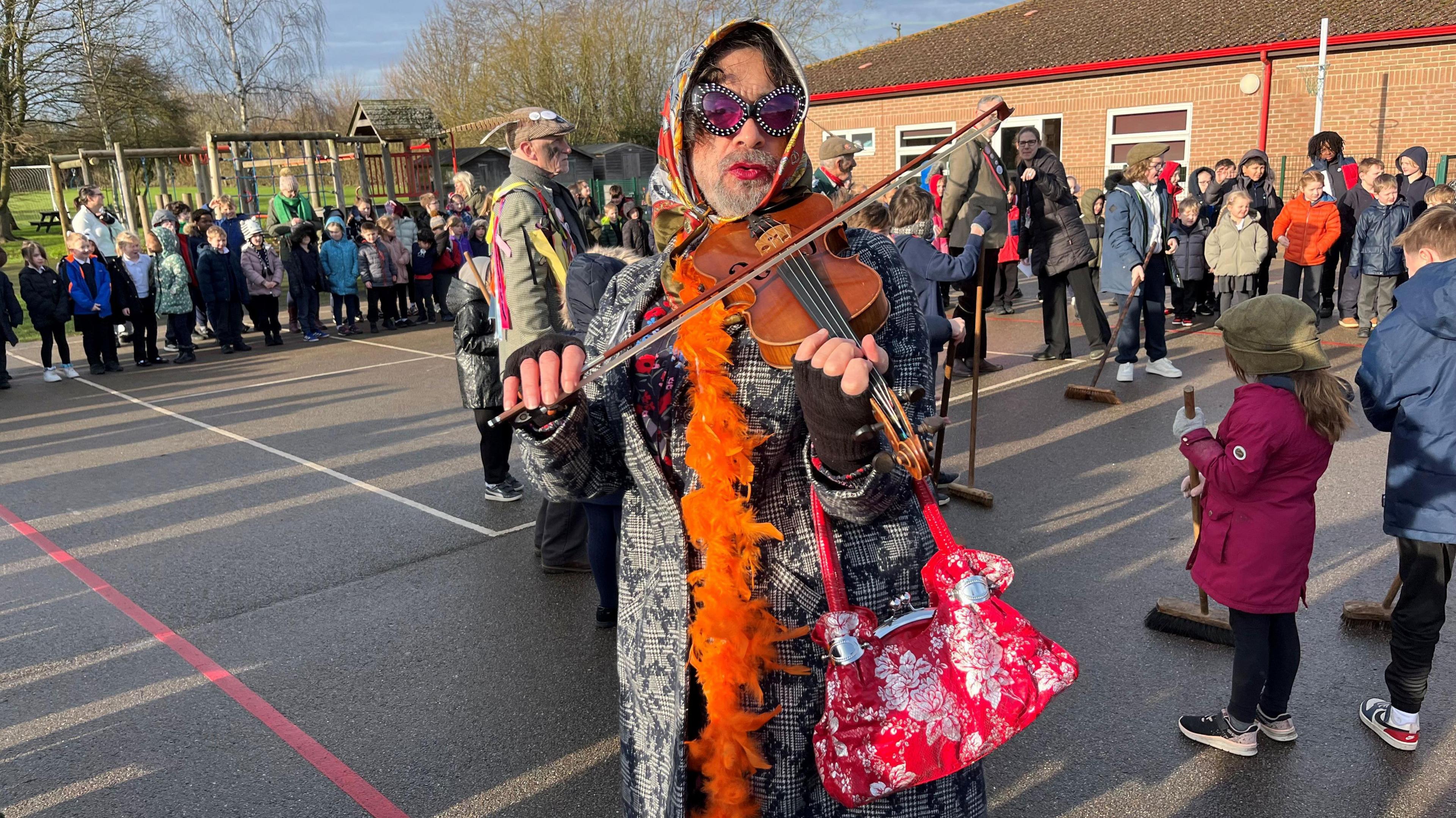 A man with a short clipped grey beard with red lipstick, oval sun glasses, a headscarf and black and white overcoat with orange feather boa plays a violin with children and fellow performers behind him.