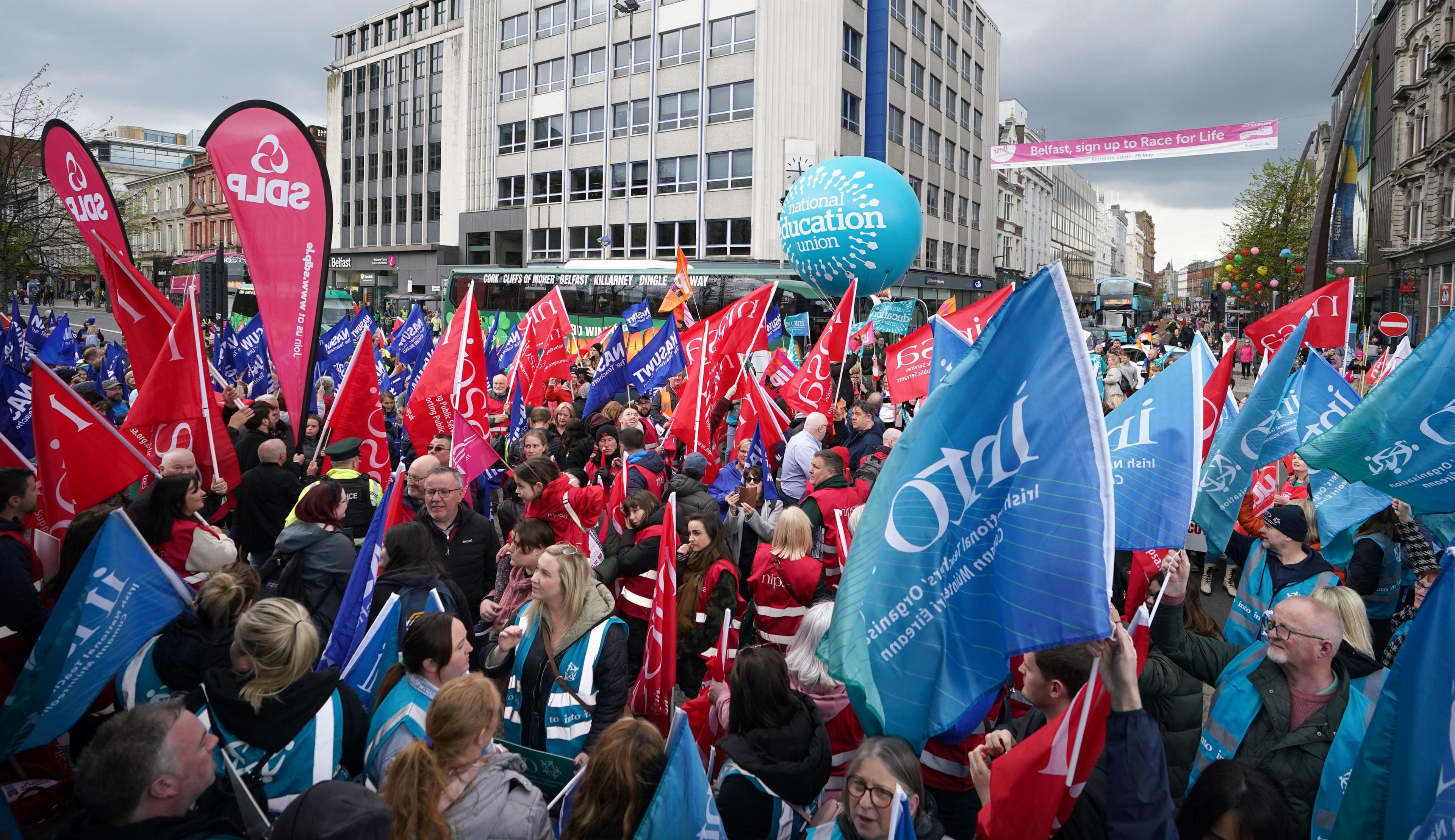 A crowd of people strike outside Belfast City Hall, some are holding red others are holding blue flags. They are also wearing blue and red high vis jackets. In the background there is a large white building and a green bus.