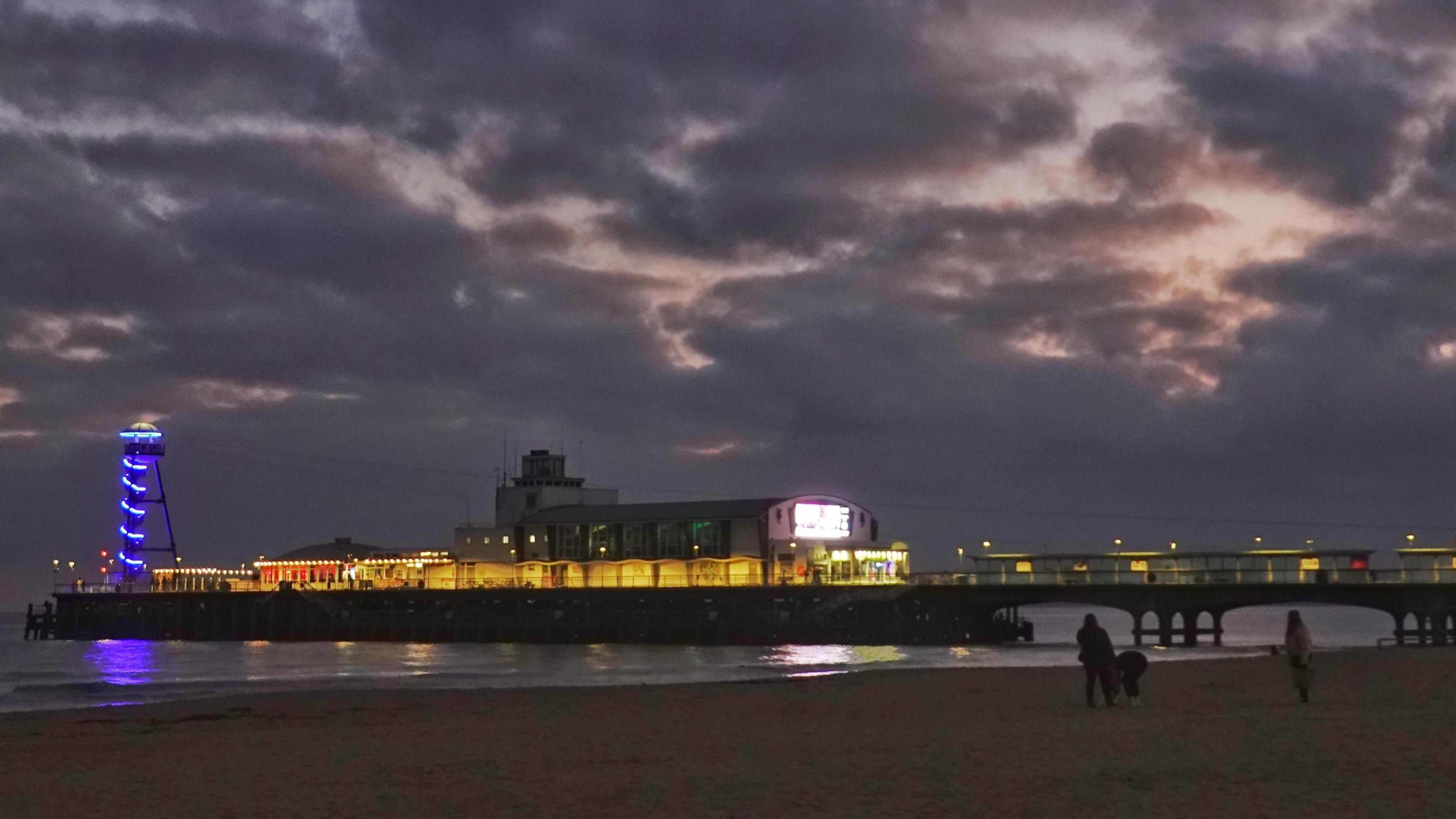 Dark clouds dominate the dusk sky which hides shades of pinks. Bournemouth pier is illuminated and  crosses the bottom third of the picture with the blue lights of a tower at its end. In the foreground some dog walkers can be seen on the beach.