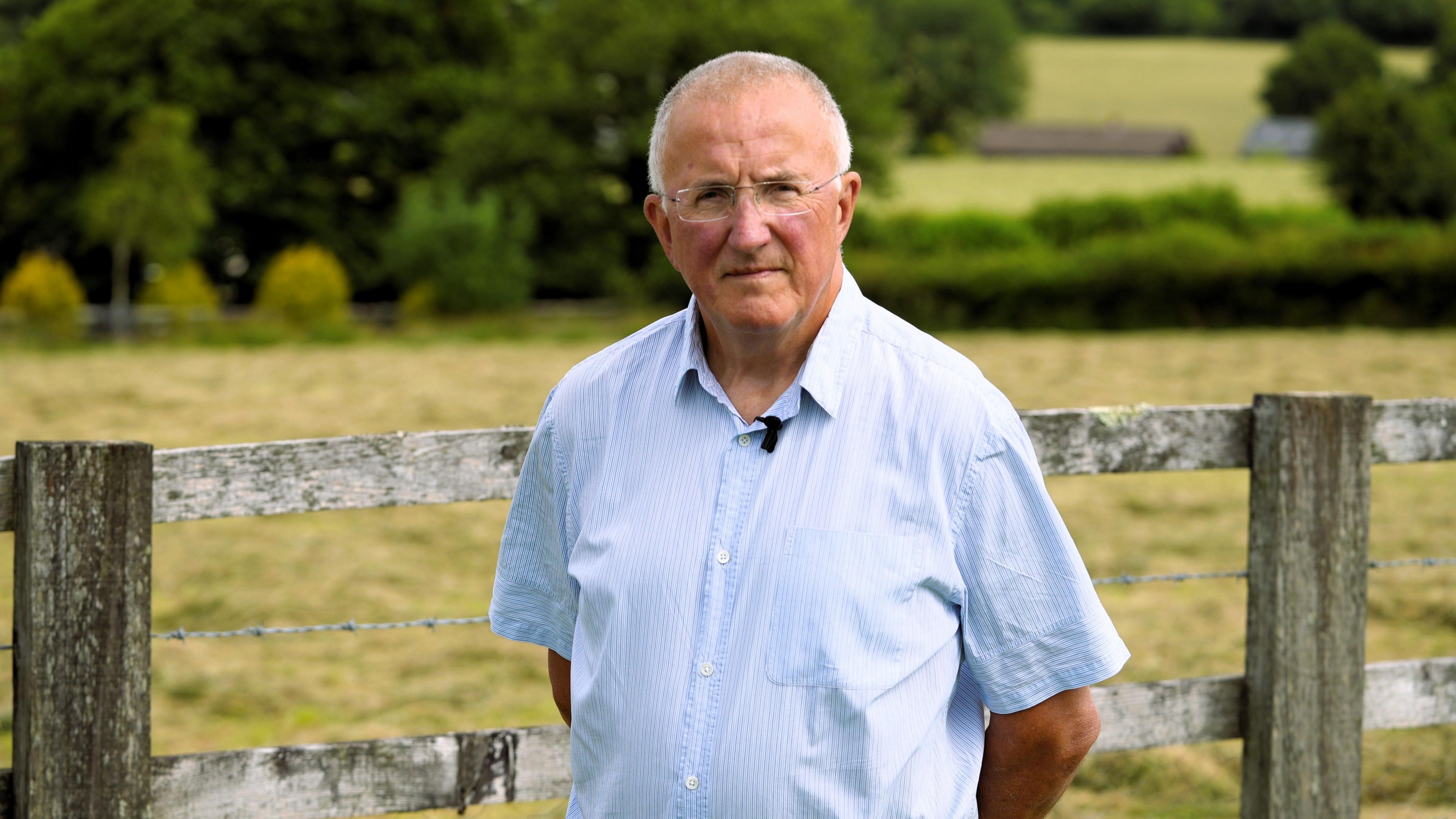 Rhodri Williams, dyn a gwallt llwyd mewn crys, yn sefyll o flaen ffens yng nghefn gwlad Cymru.