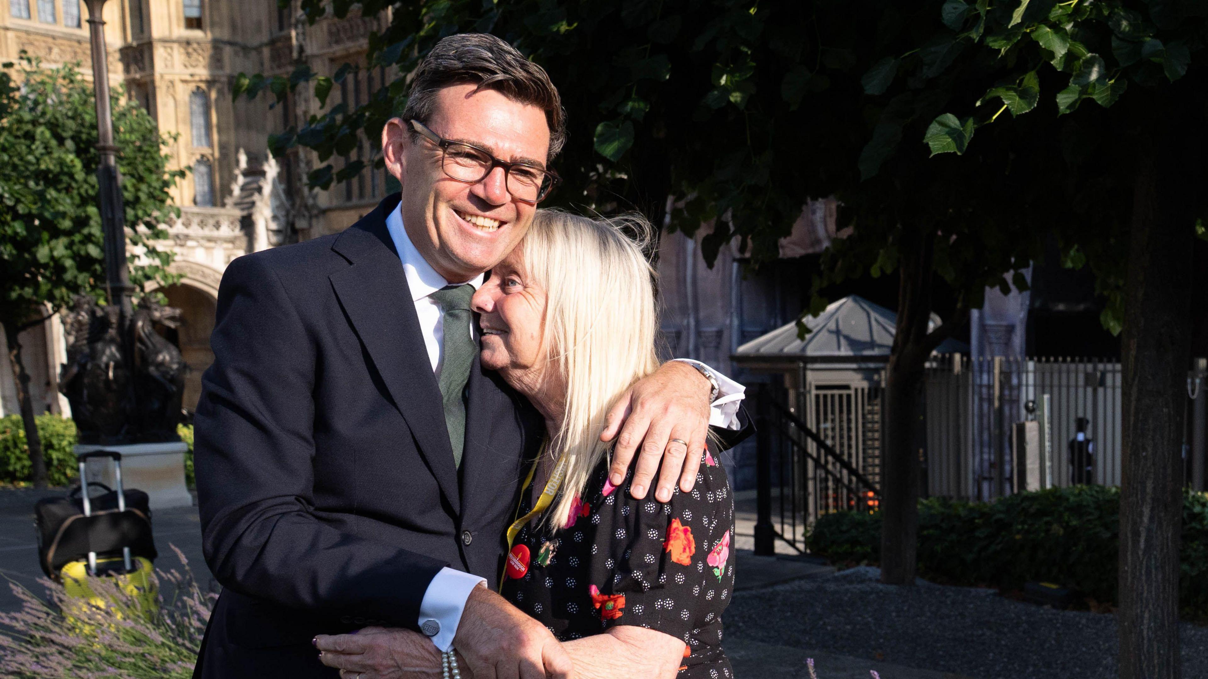 A man in a dark suit, wearing glasses and smiling broadly hugs a blond woman in a black floral dress on a sunny day outside the houses of parliament. 