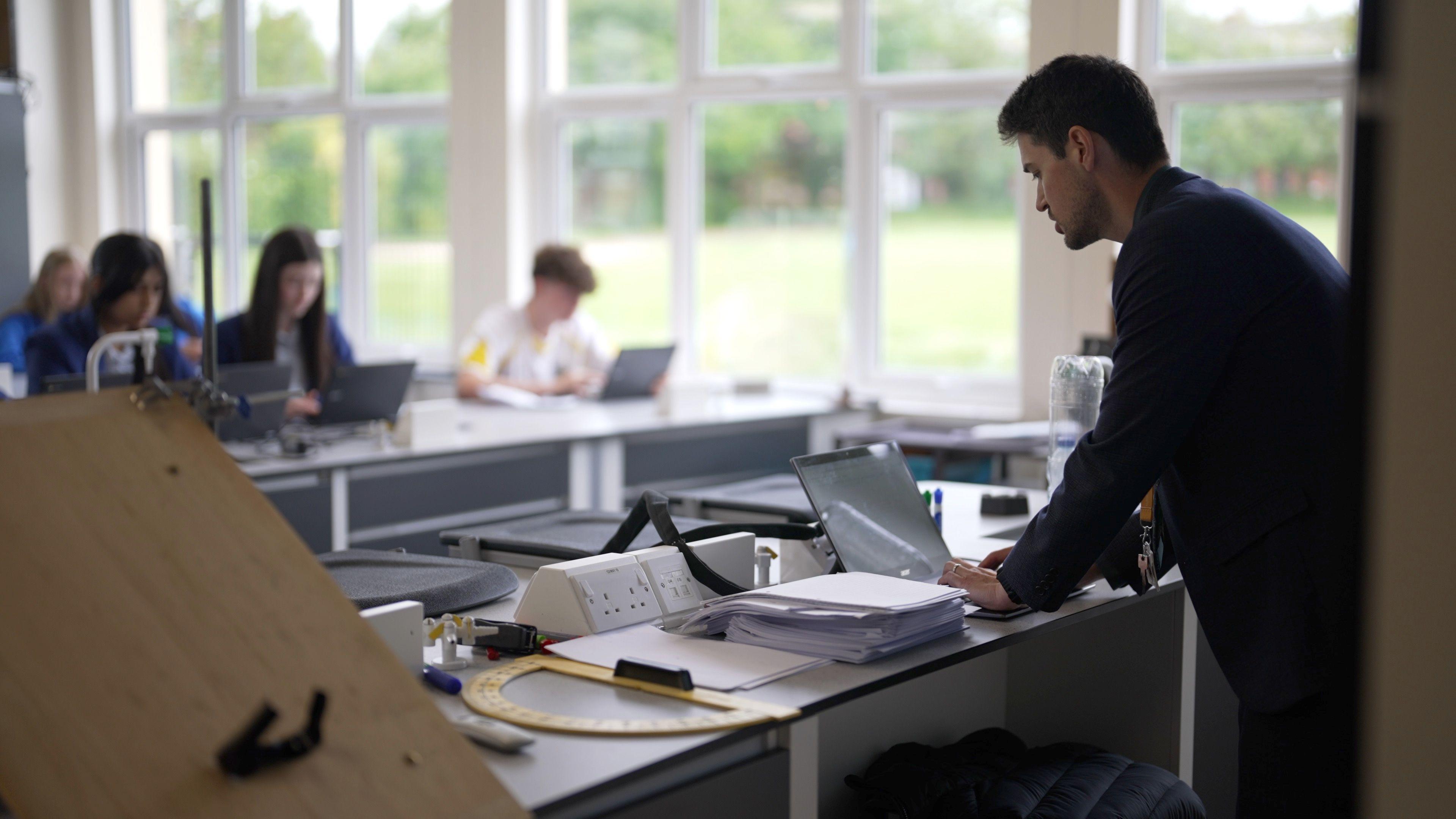 A teacher in a black top looks down at his laptop at the top of the classroom. There are students sitting on rows of desk listening - all with laptops. 