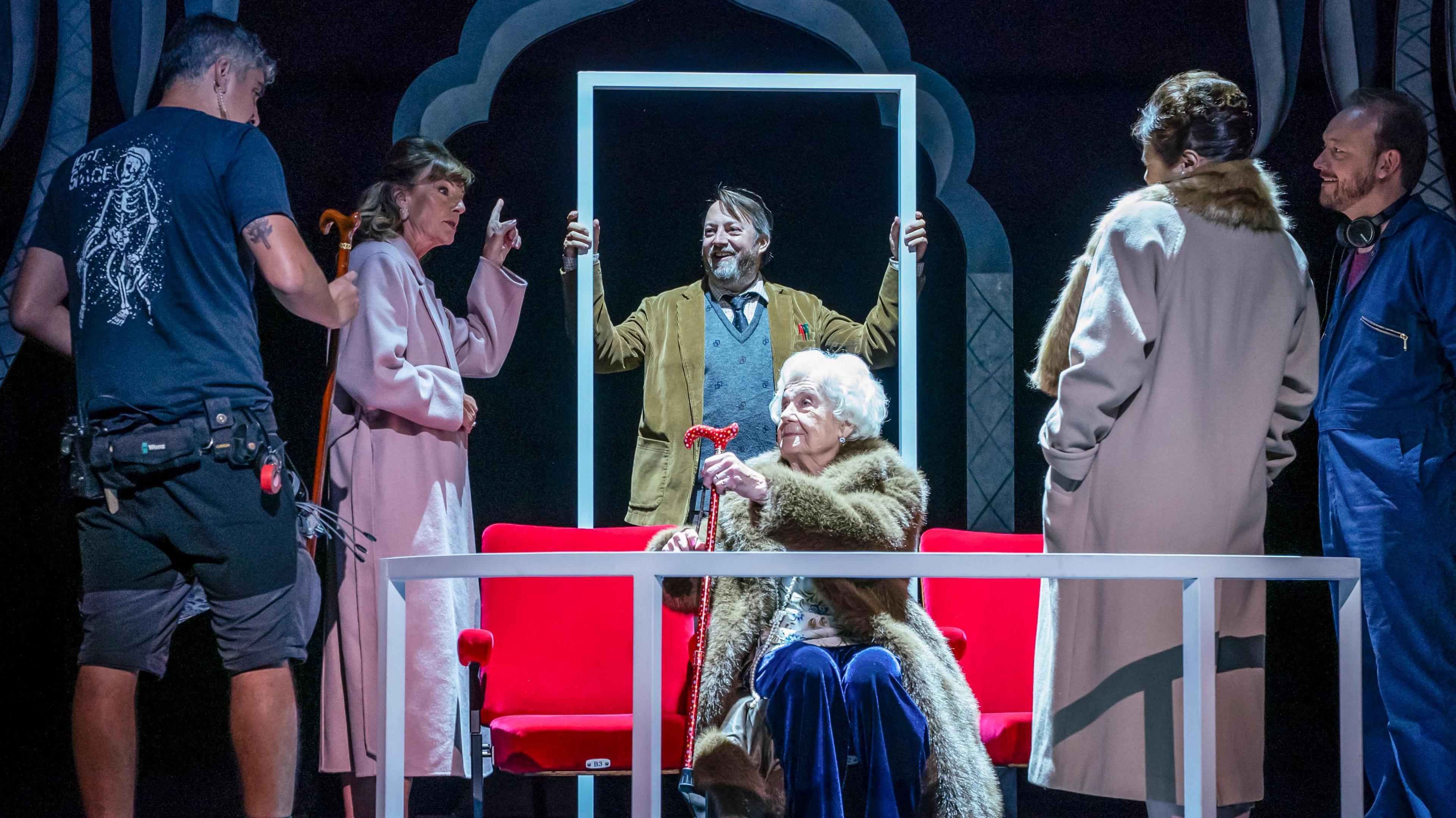 David Mitchell in a brown blazer and grey jumper stands in the centre of the stage, in front of him, sitting on a red sofa is an elderly woman in a fur coat. In either side of the photo there are two women wearing pink coats and next to them camera men dressed in blue.