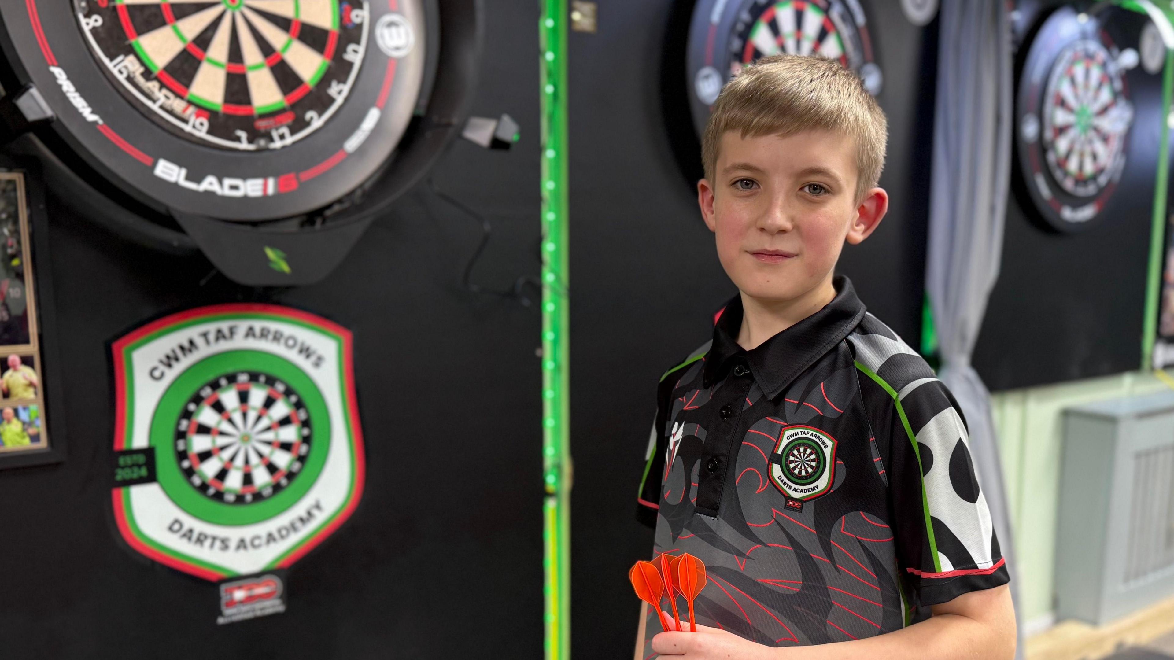 Jac, 12, stands next to a dartboard at Cwm Taf Arrows Darts Academy and holds his darts