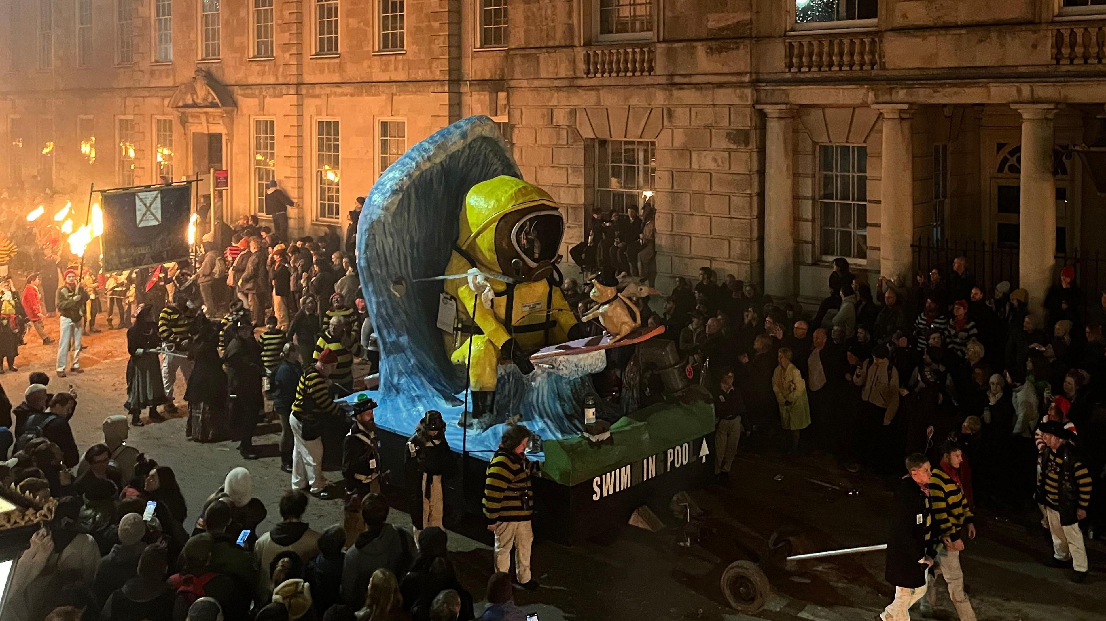 People stood sround an effigy of a yellow hazmat suit sat in a wave with the words SWIM IN POO on the front of the float. People are holding fire torches at the back of the effigy