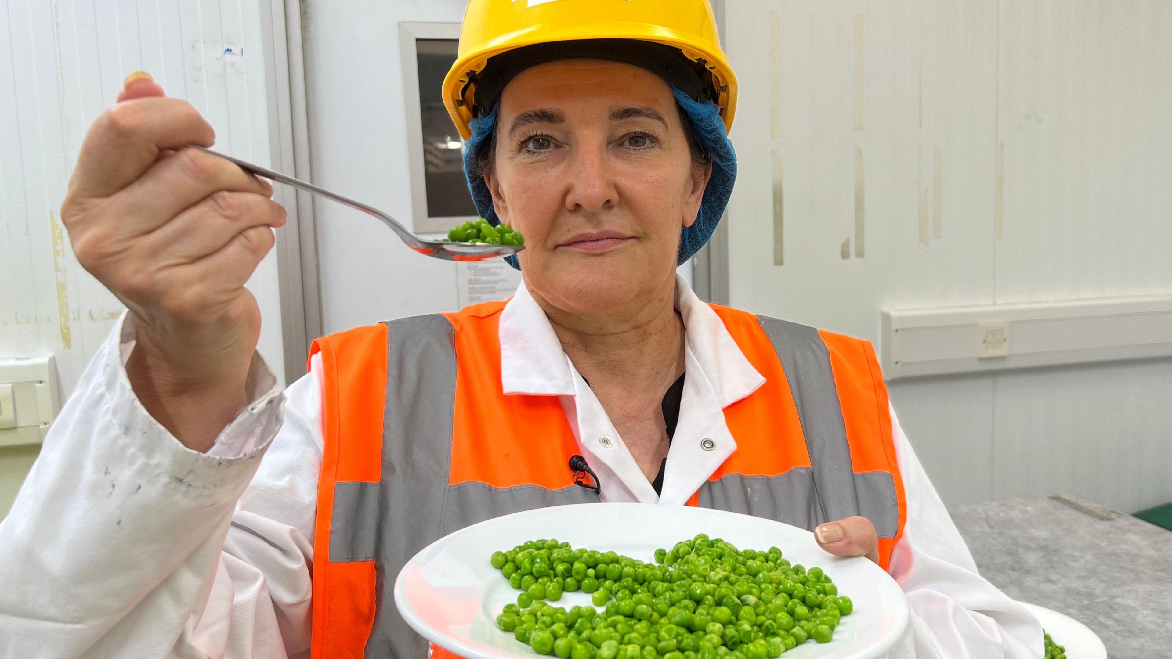 Michelle is wearing an orange high-vis vest over a white lab coat. She has a yellow hard hat on her head and a blue net covering her hair. She is holding a plate of peas in the right hand and a spoon full of peas with the left hand. The spoon is being held up to her face.
