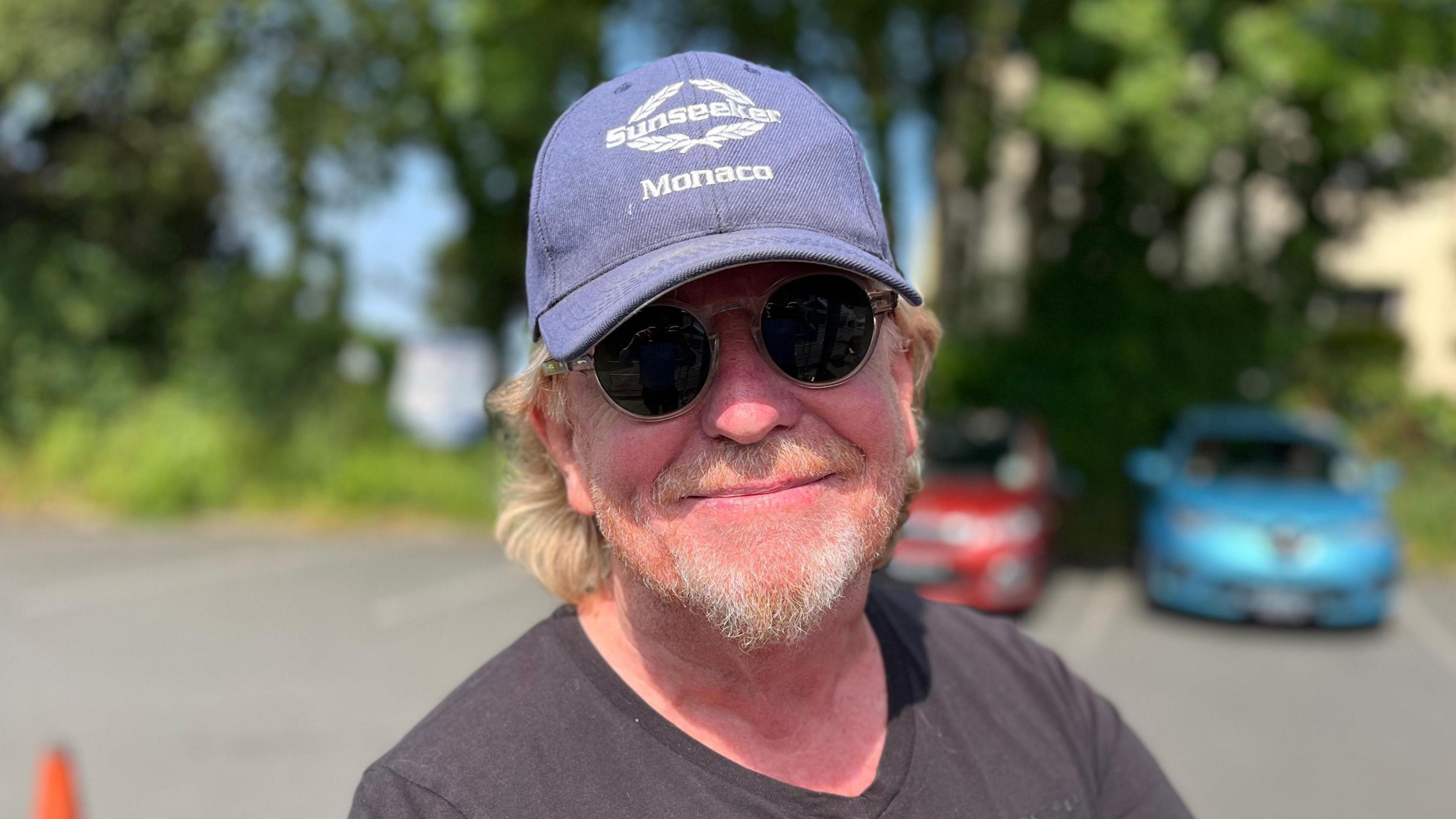 Deputy Bopb Murray wearing a blue hat and black sunglasses and smiling into the camera. He has long ginger hair and a ginger and grey beard.