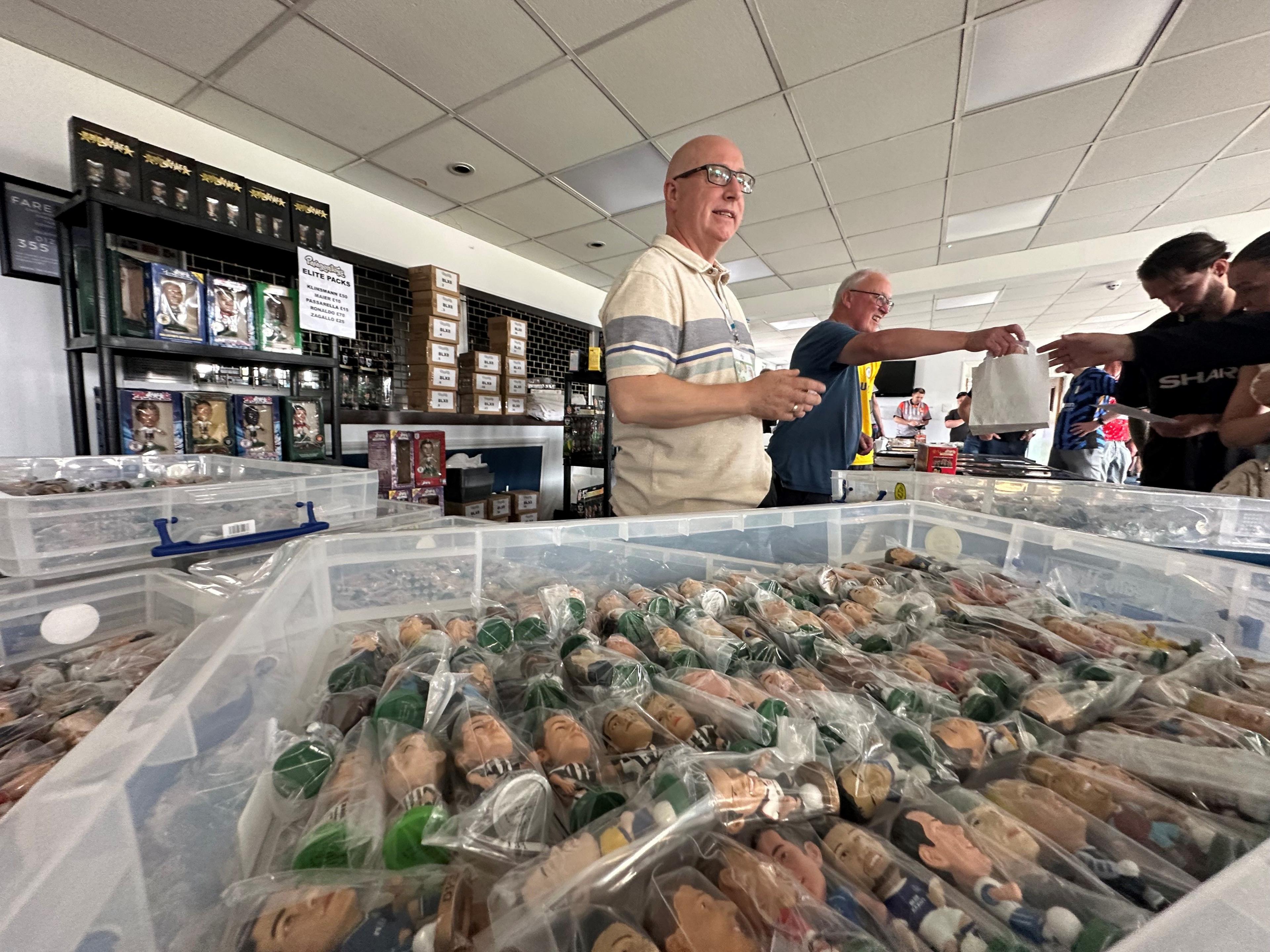 Several people look at trays of football figures on a table, behind the table are two dealers talking to buyers, one of whom is handing over a white paper bag.